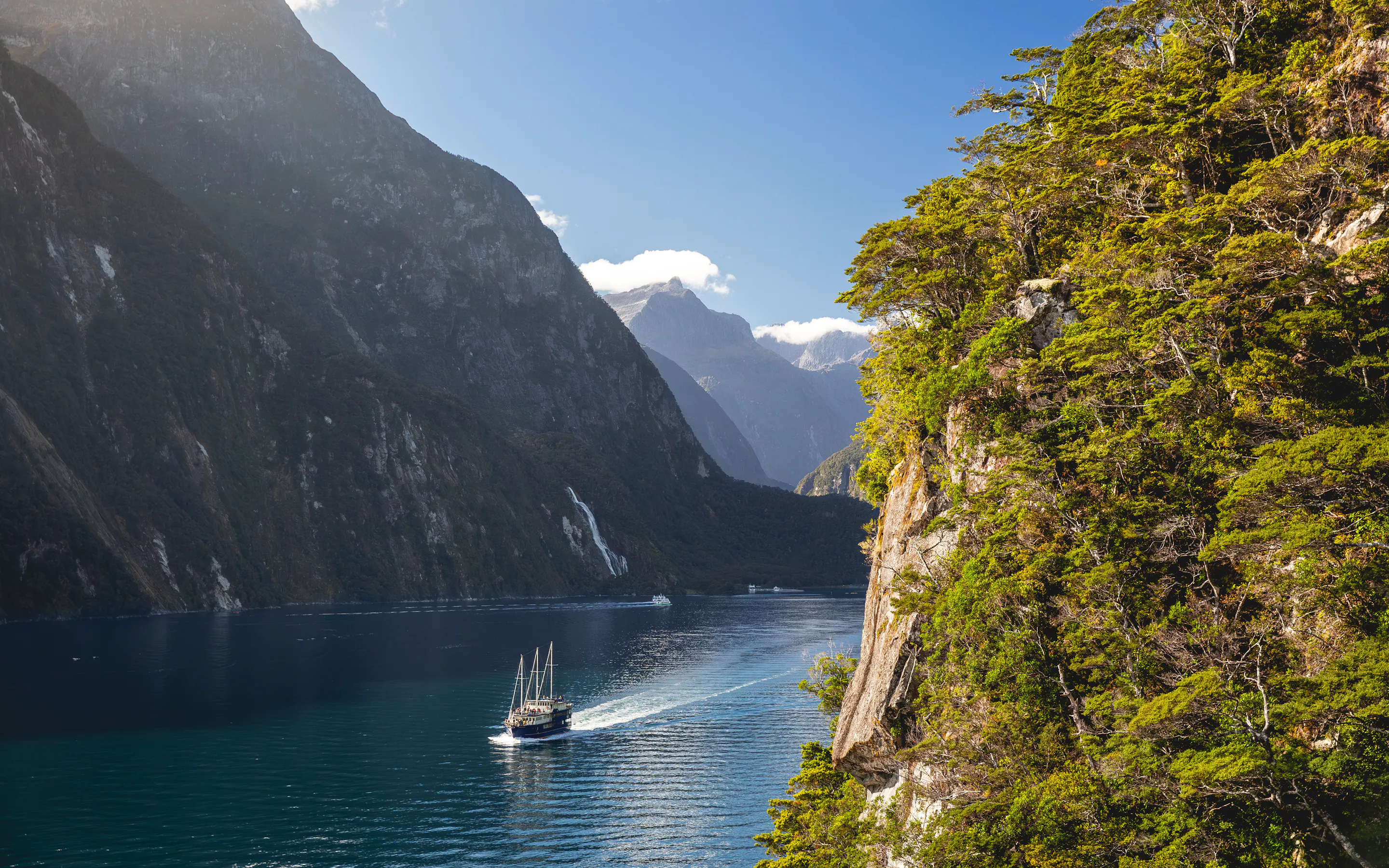 A small boat glides across dark water beneath sheer fiord cliffs, with low cloud clinging to the mountain slopes.