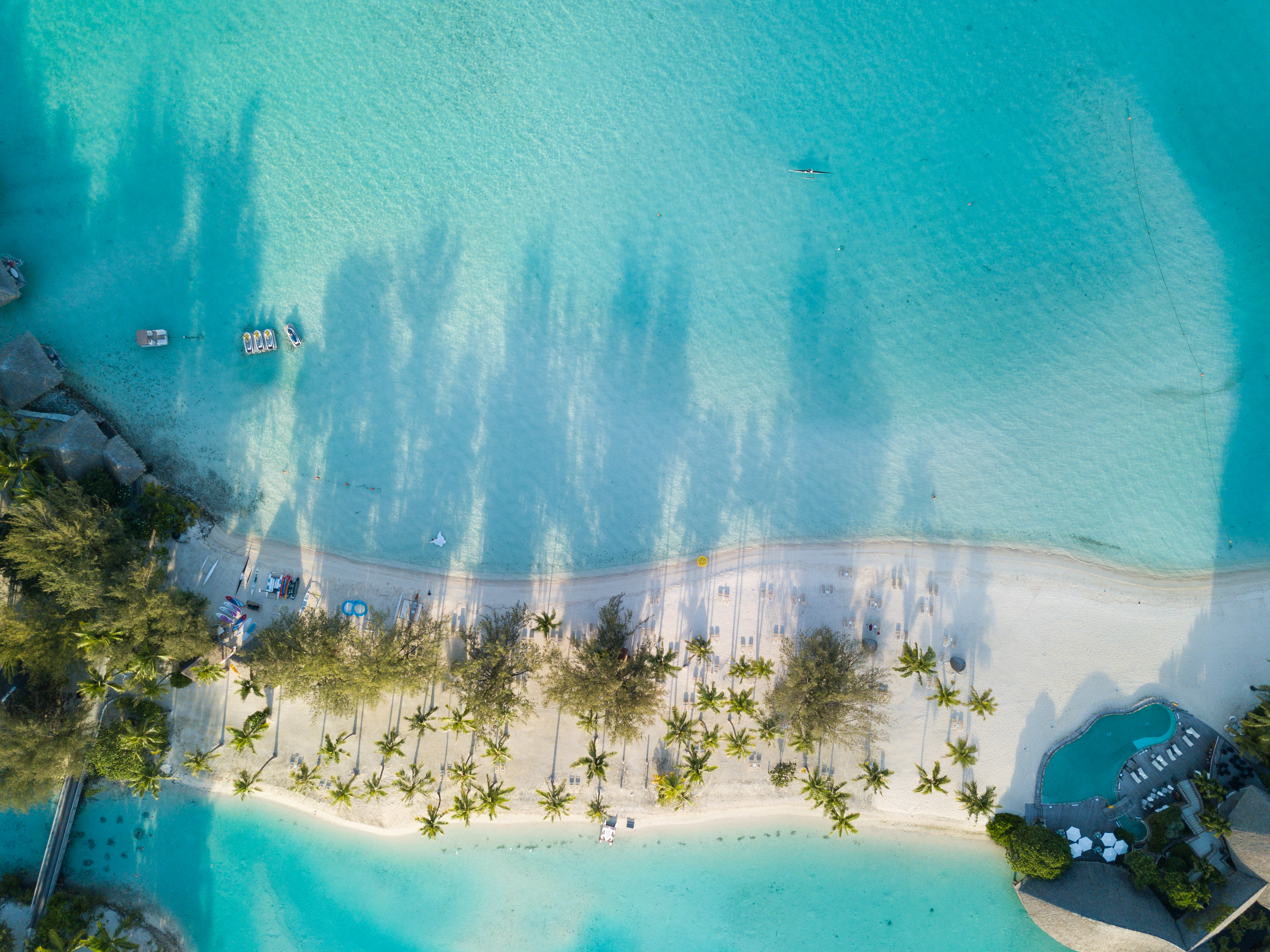An aerial view shows a pale sand beach curving into bright blue water, edged by palms and shallow coral flats.