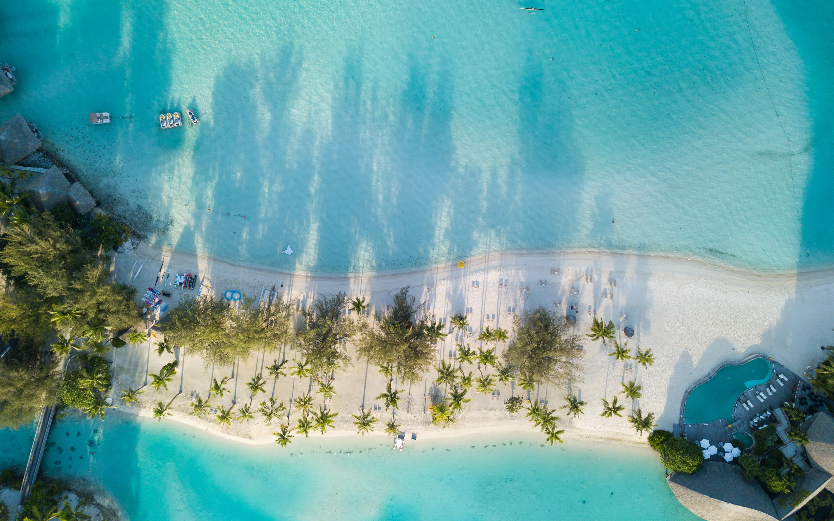 An aerial view shows a pale sand beach curving into bright blue water, edged by palms and shallow coral flats.