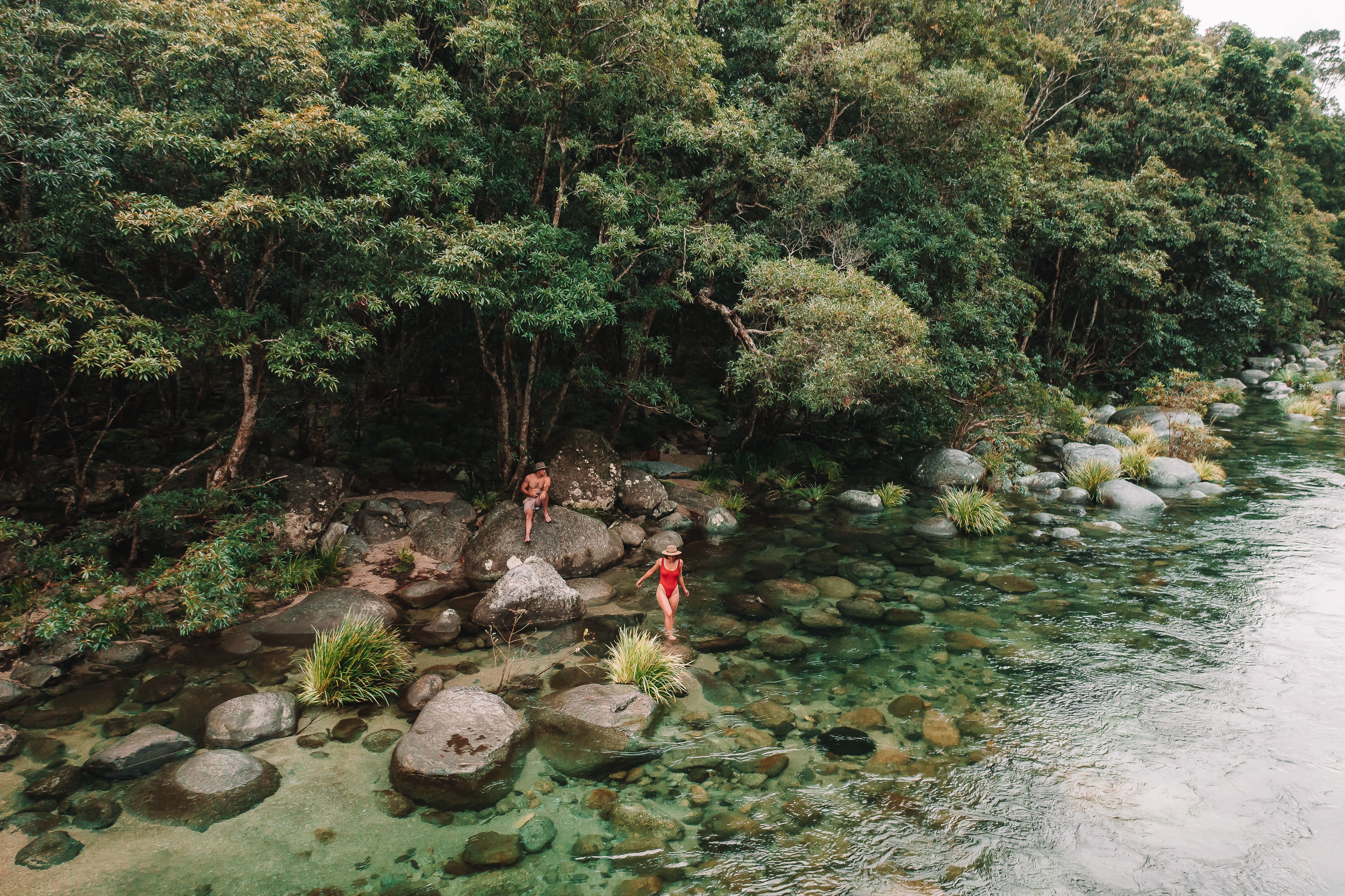Two adults wade among smooth river boulders beside dense rainforest, with clear green water flowing past the shore.