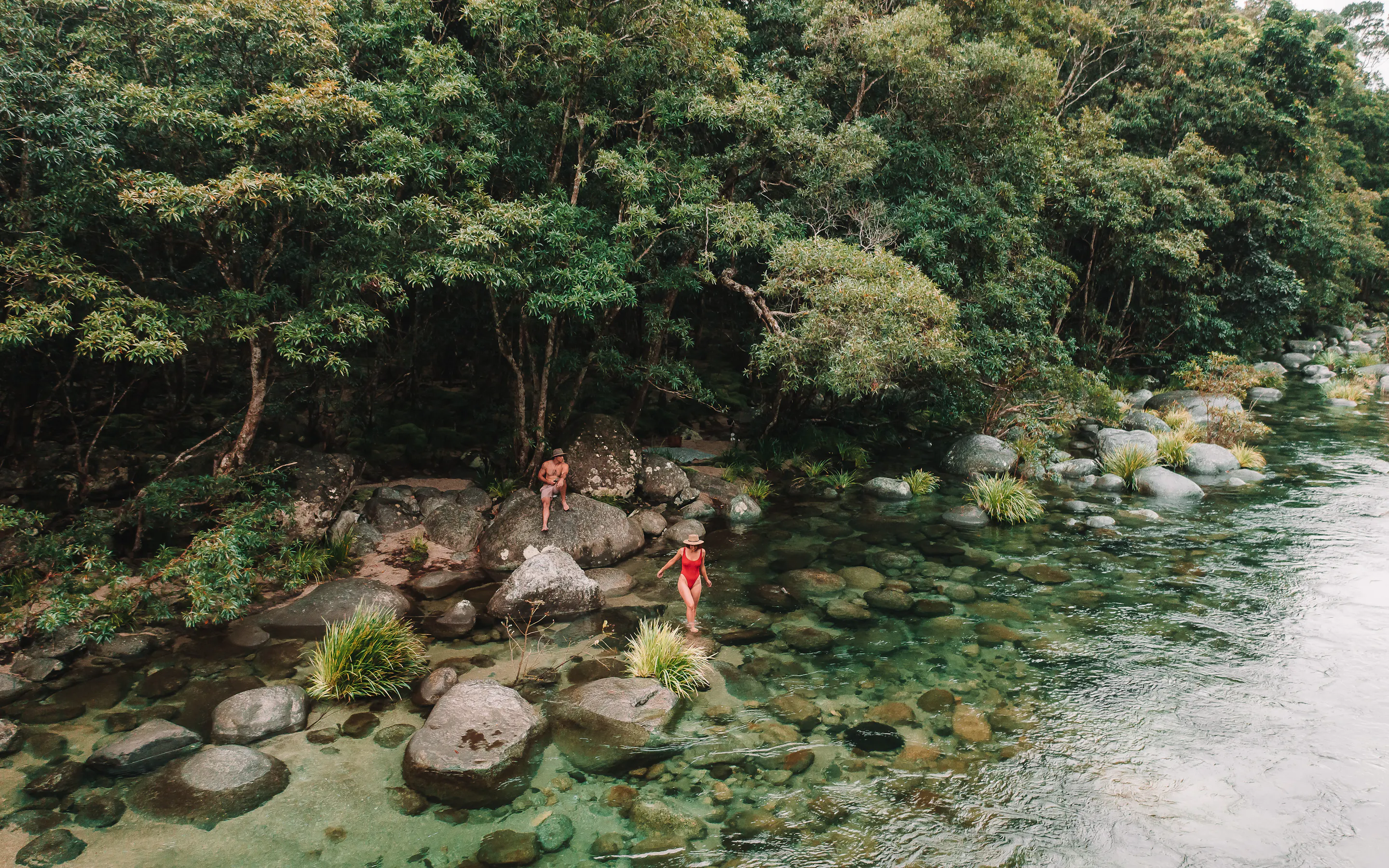 Two adults wade among smooth river boulders beside dense rainforest, with clear green water flowing past the shore.