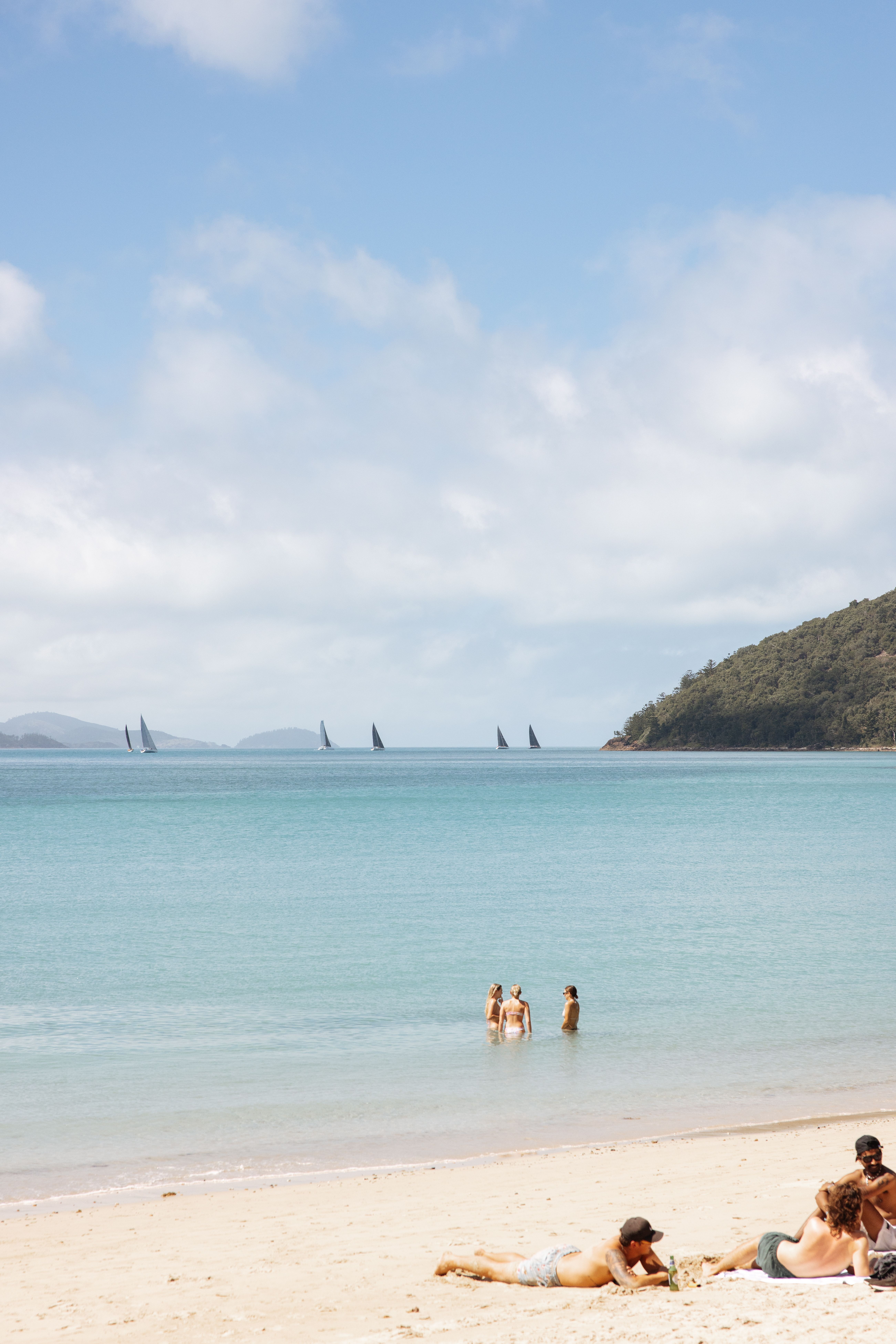 Three people wade in a calm bay near a sandy beach, with sailboats and a low forested island in the distance.