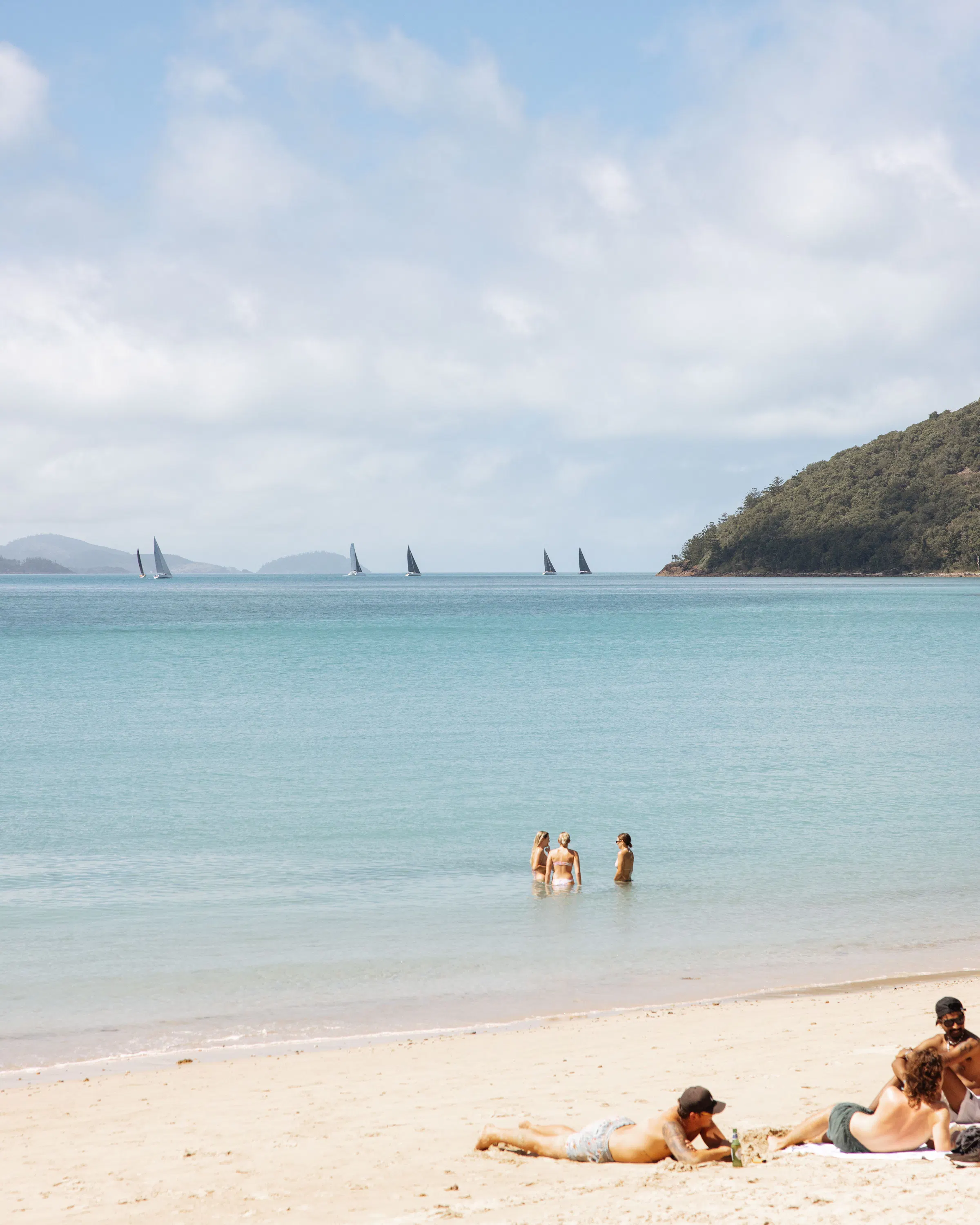 Three people wade in a calm bay near a sandy beach, with sailboats and a low forested island in the distance.