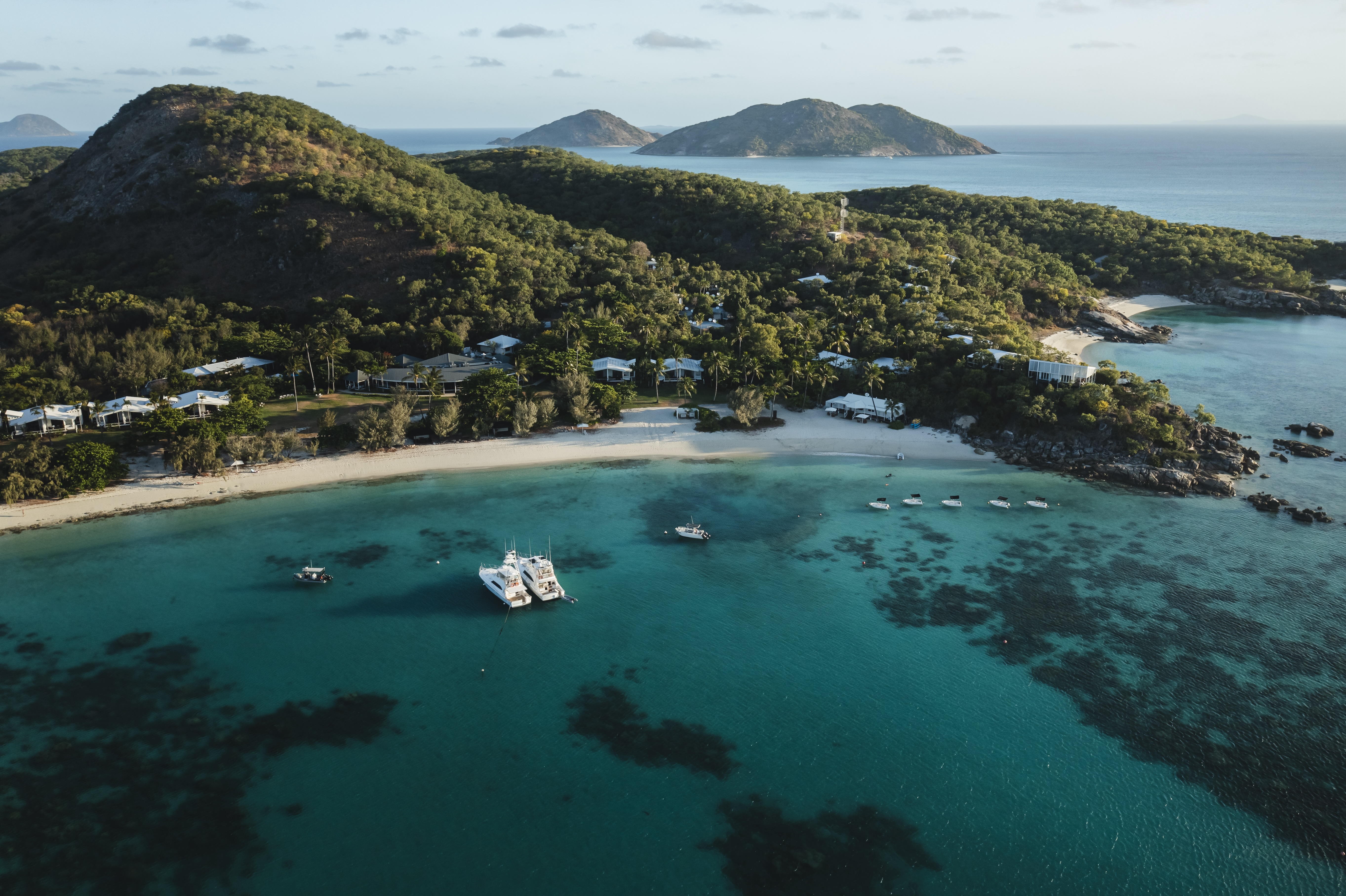 An aerial view shows a beachfront resort beside bright reef water, with moored boats and wooded hills beyond.