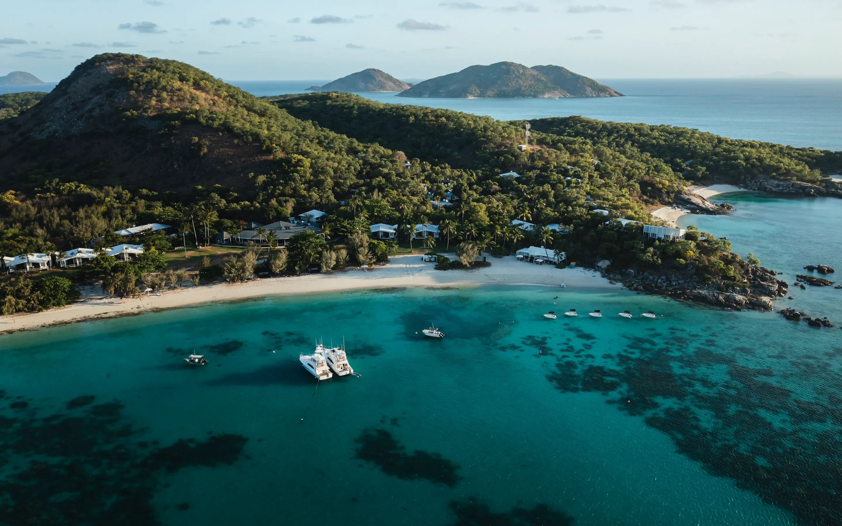 An aerial view shows a beachfront resort beside bright reef water, with moored boats and wooded hills beyond.