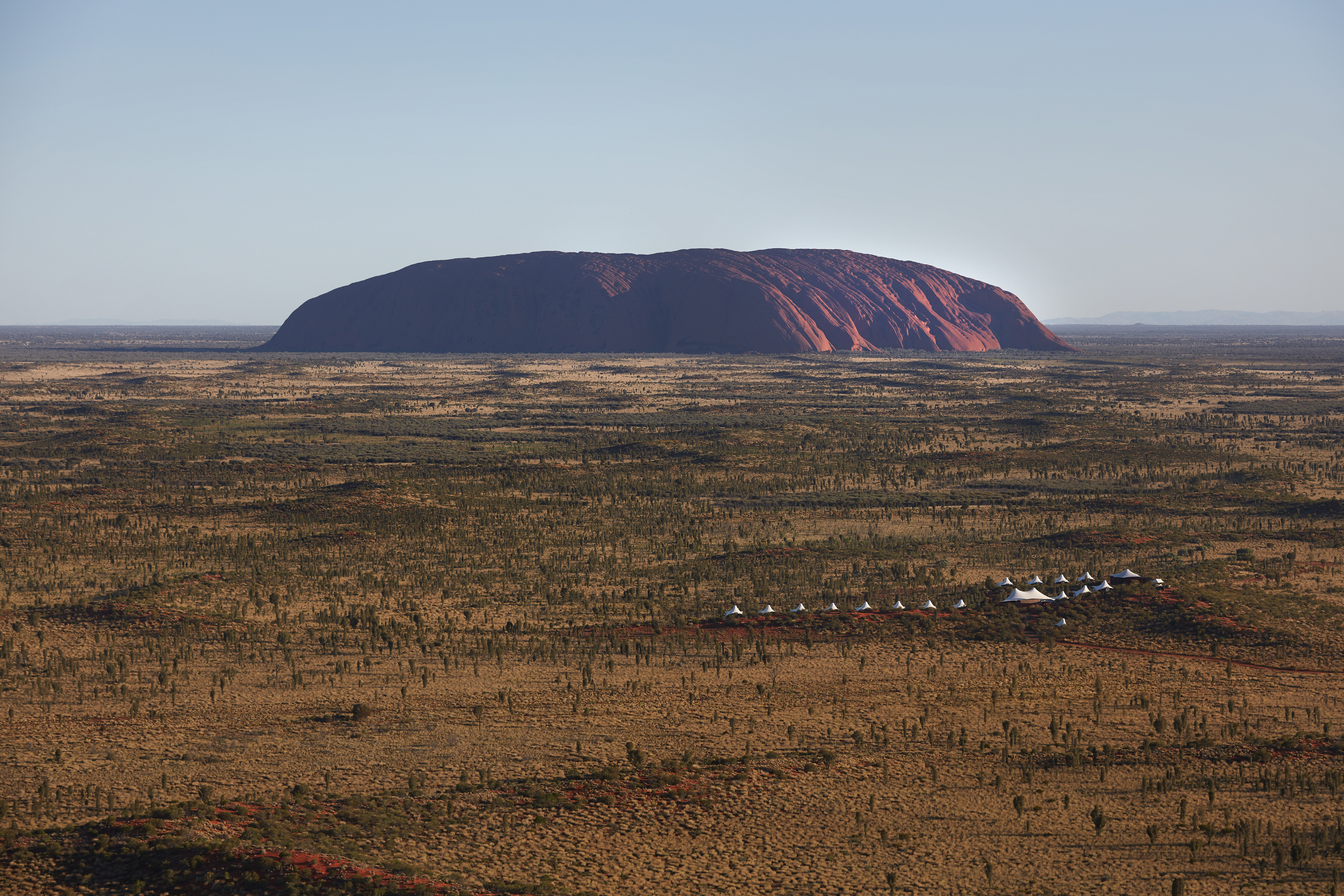 An aerial view shows white lodge tents below Uluru, with ocher earth and low desert scrub spreading outward.