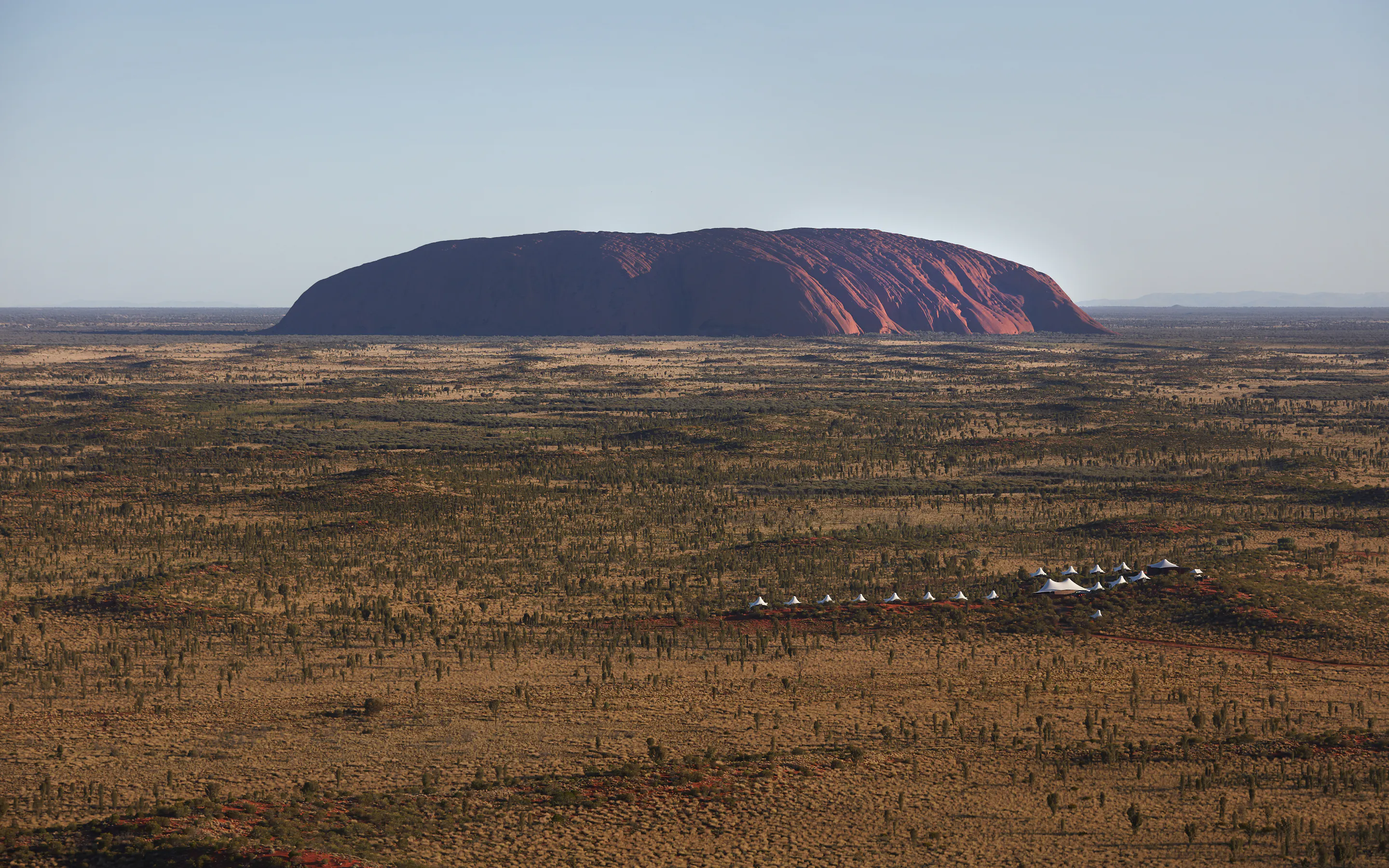 An aerial view shows white lodge tents below Uluru, with ocher earth and low desert scrub spreading outward.