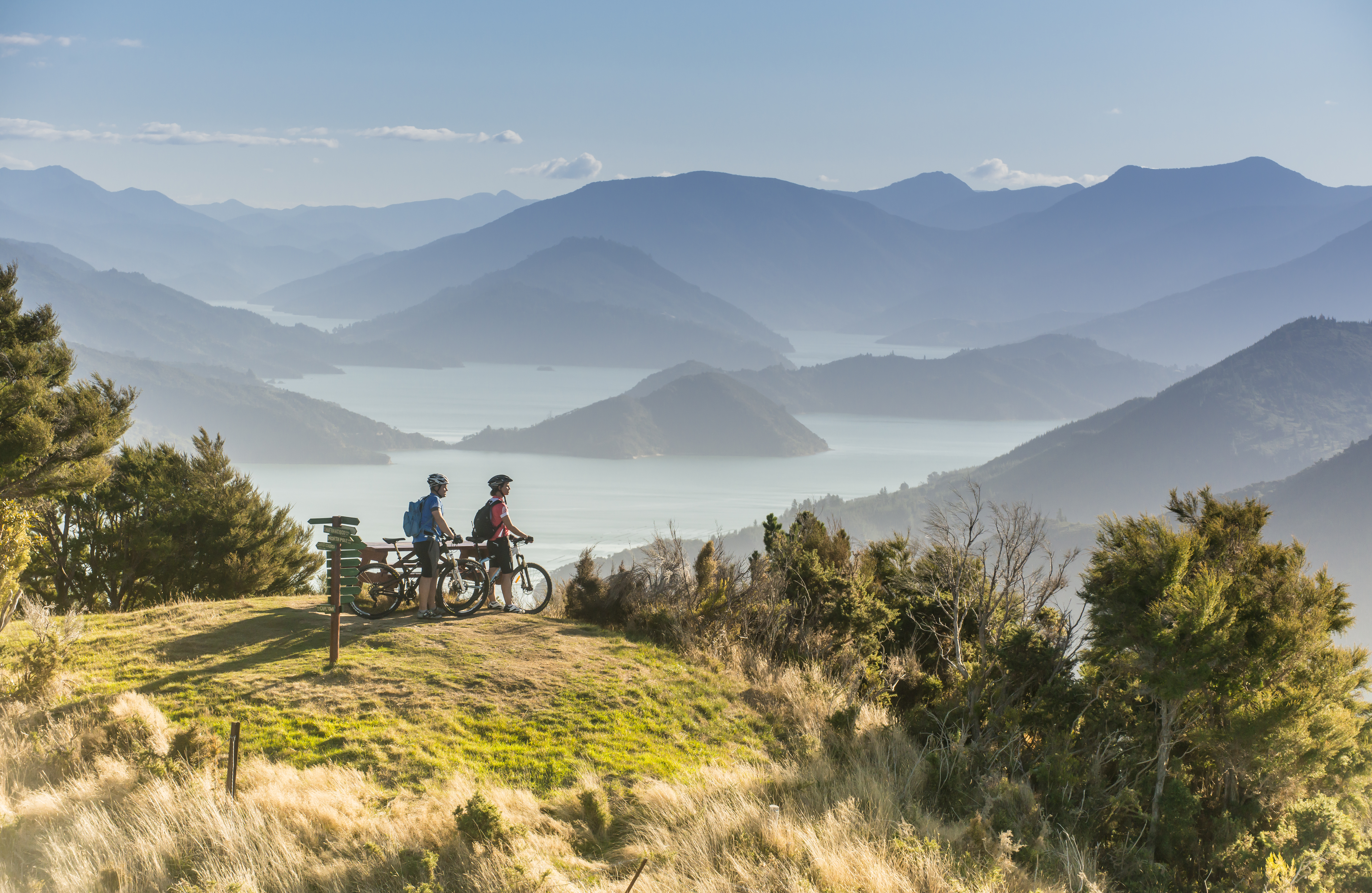 Two cyclists pause at a grassy overlook above the Marlborough Sounds, with sunlit ridges and blue inlets beyond.