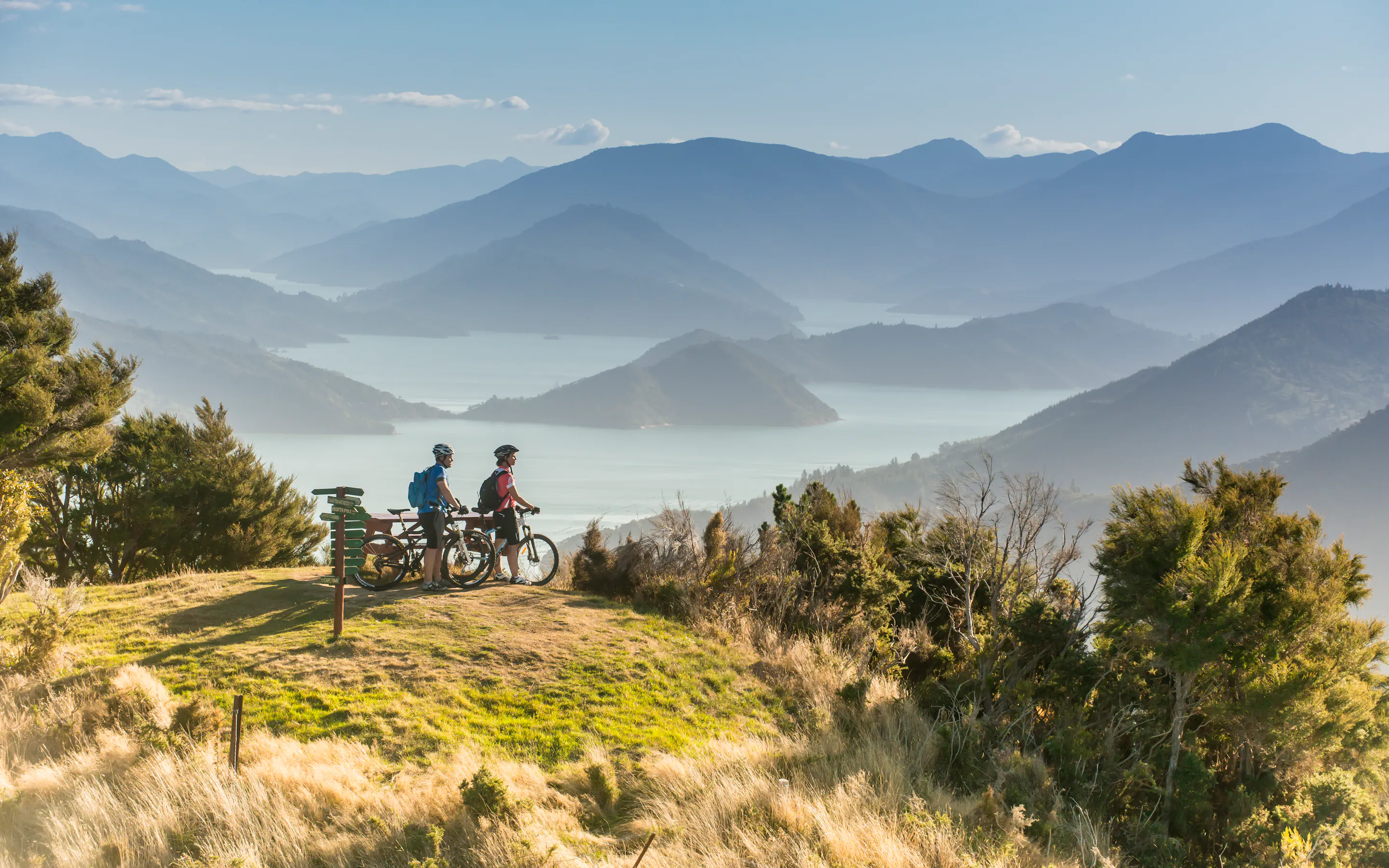 Two cyclists pause at a grassy overlook above the Marlborough Sounds, with sunlit ridges and blue inlets beyond.