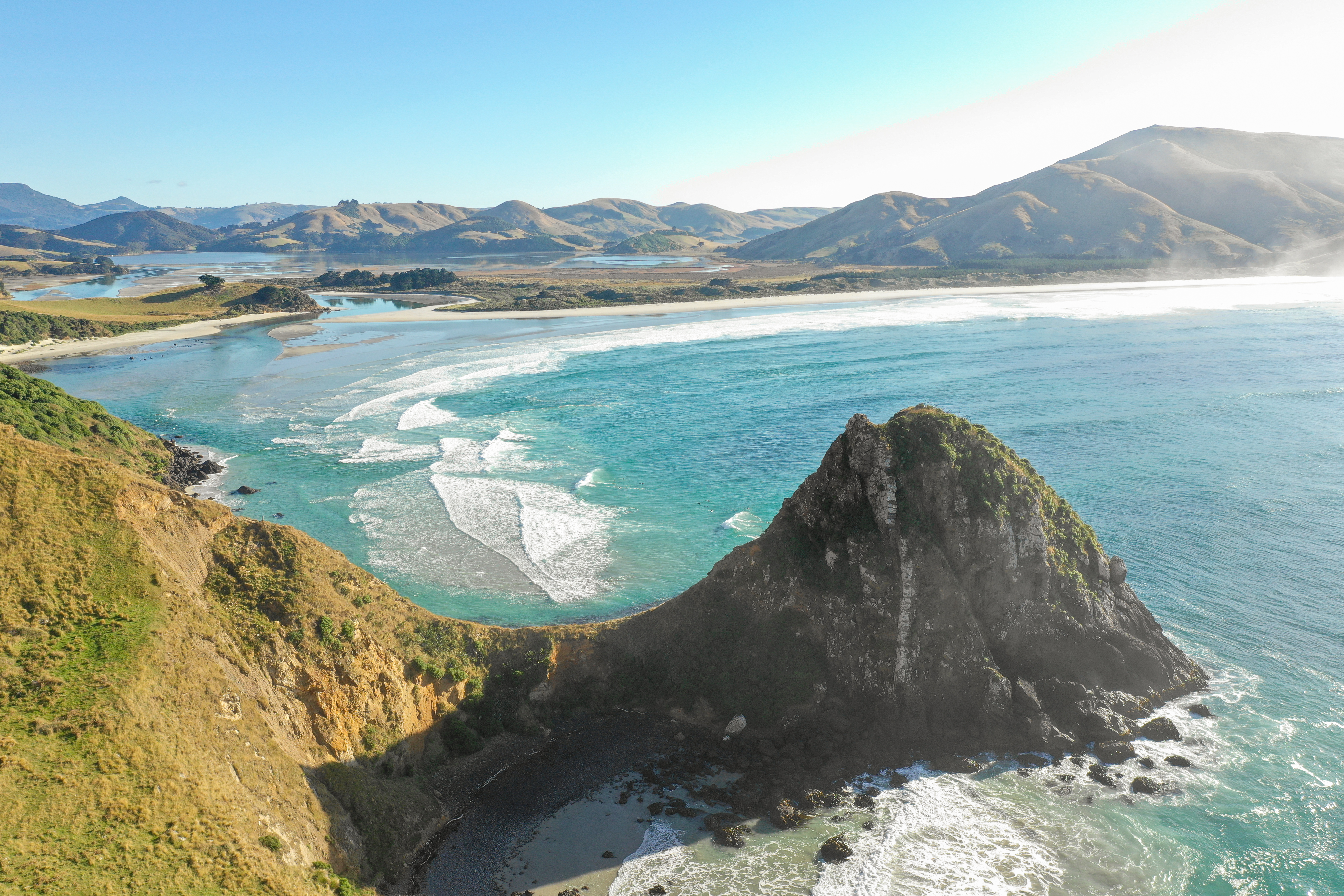 A high coastal view shows bright surf curling around a rocky headland and sweeping bay backed by sunlit hills.