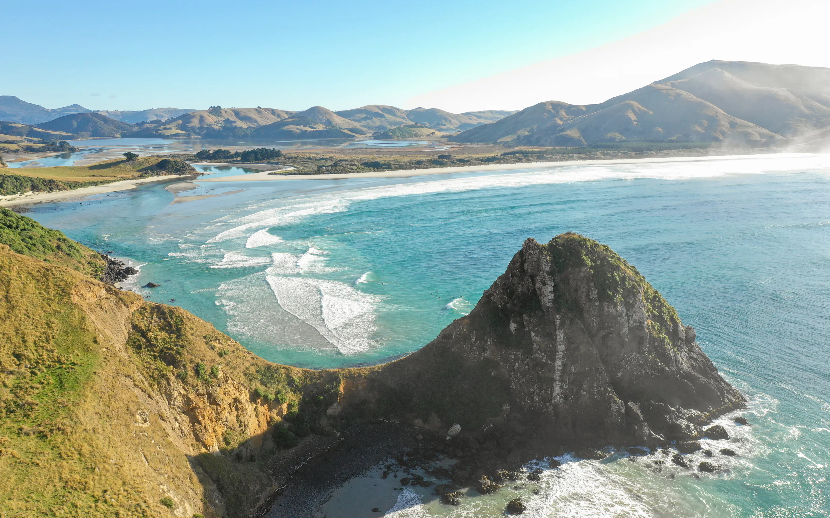 A high coastal view shows bright surf curling around a rocky headland and sweeping bay backed by sunlit hills.