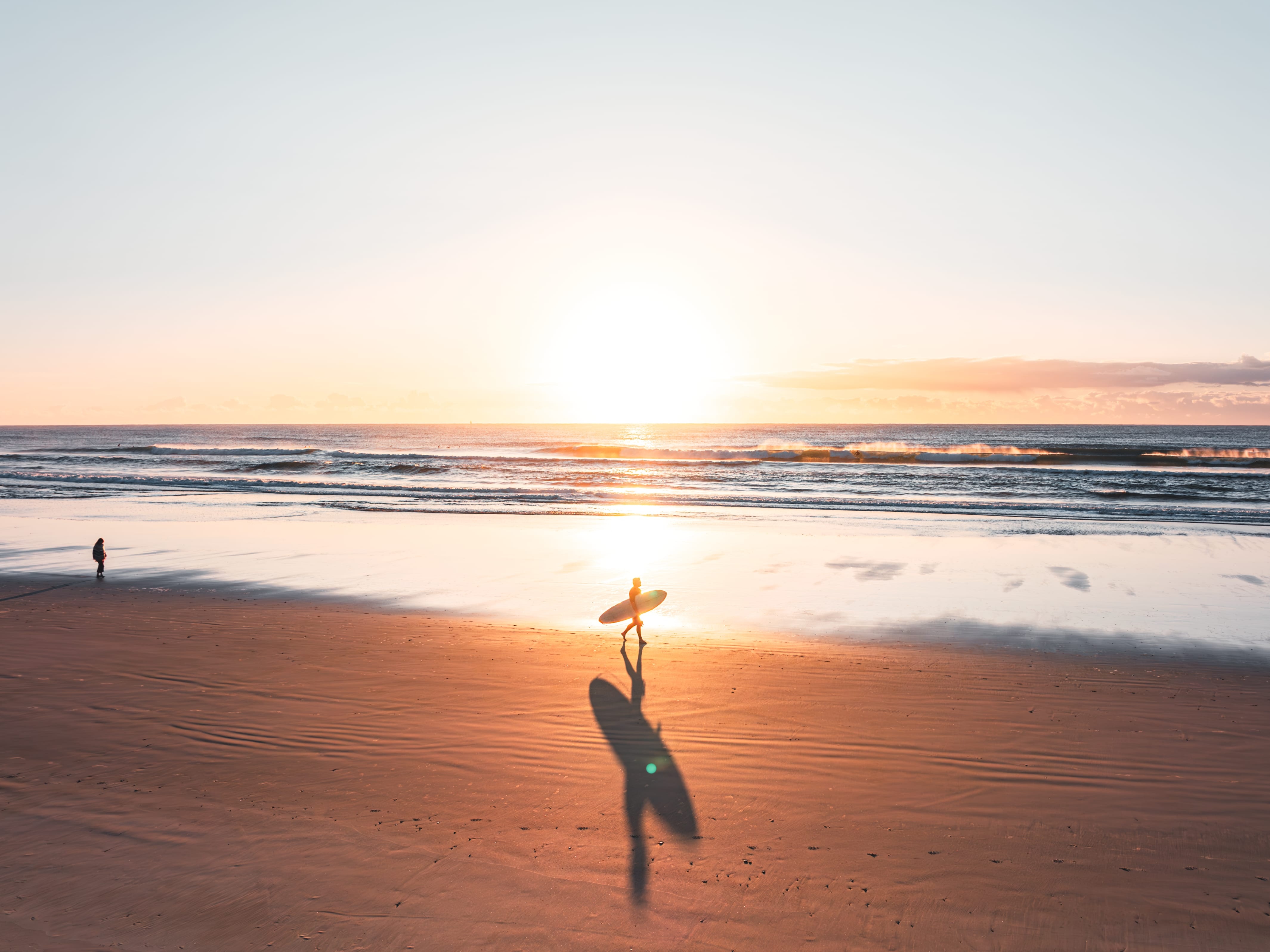 A lone surfer walks across reflective wet sand at sunset, carrying a board toward low waves and a golden sky.