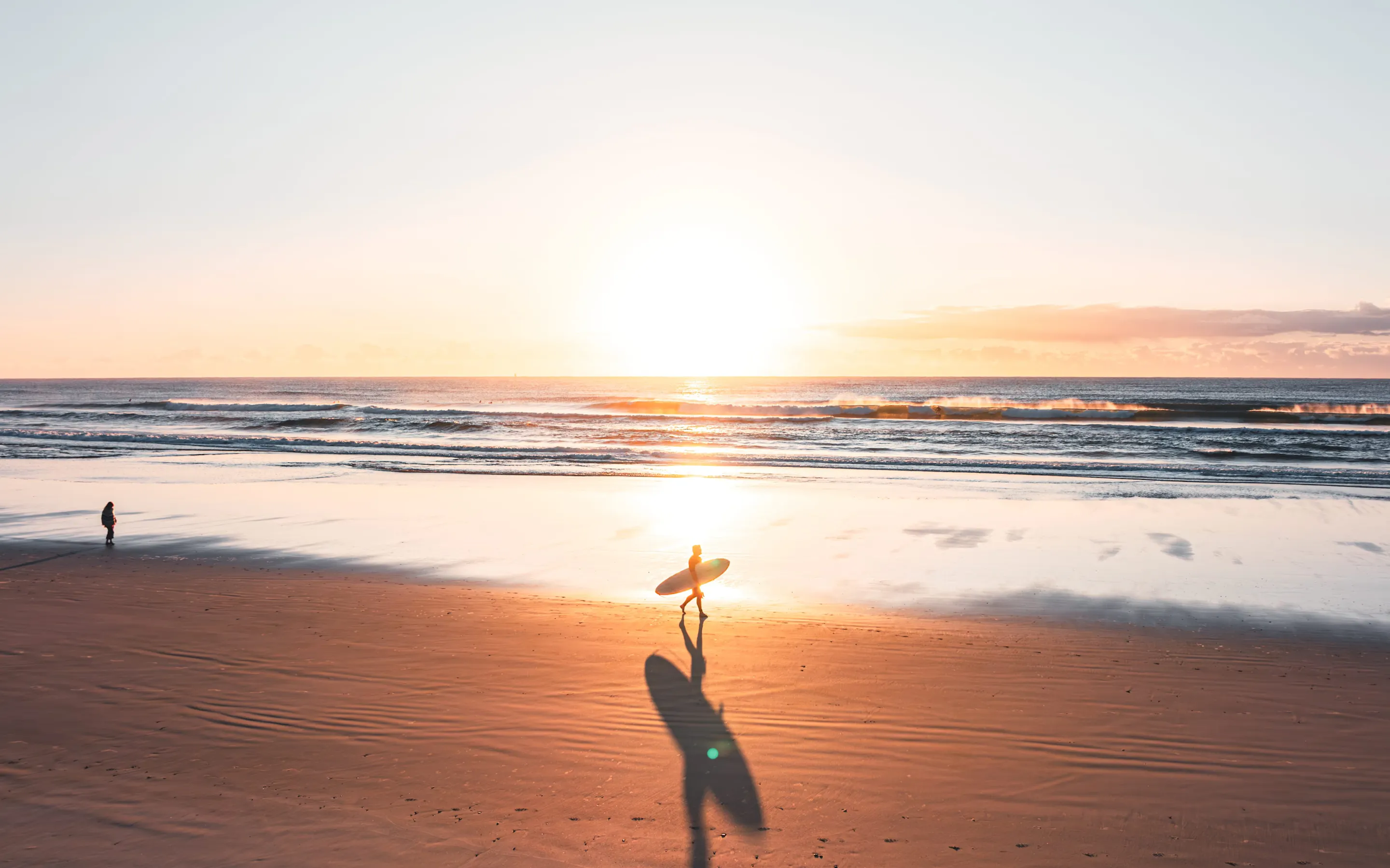 A lone surfer walks across reflective wet sand at sunset, carrying a board toward low waves and a golden sky.