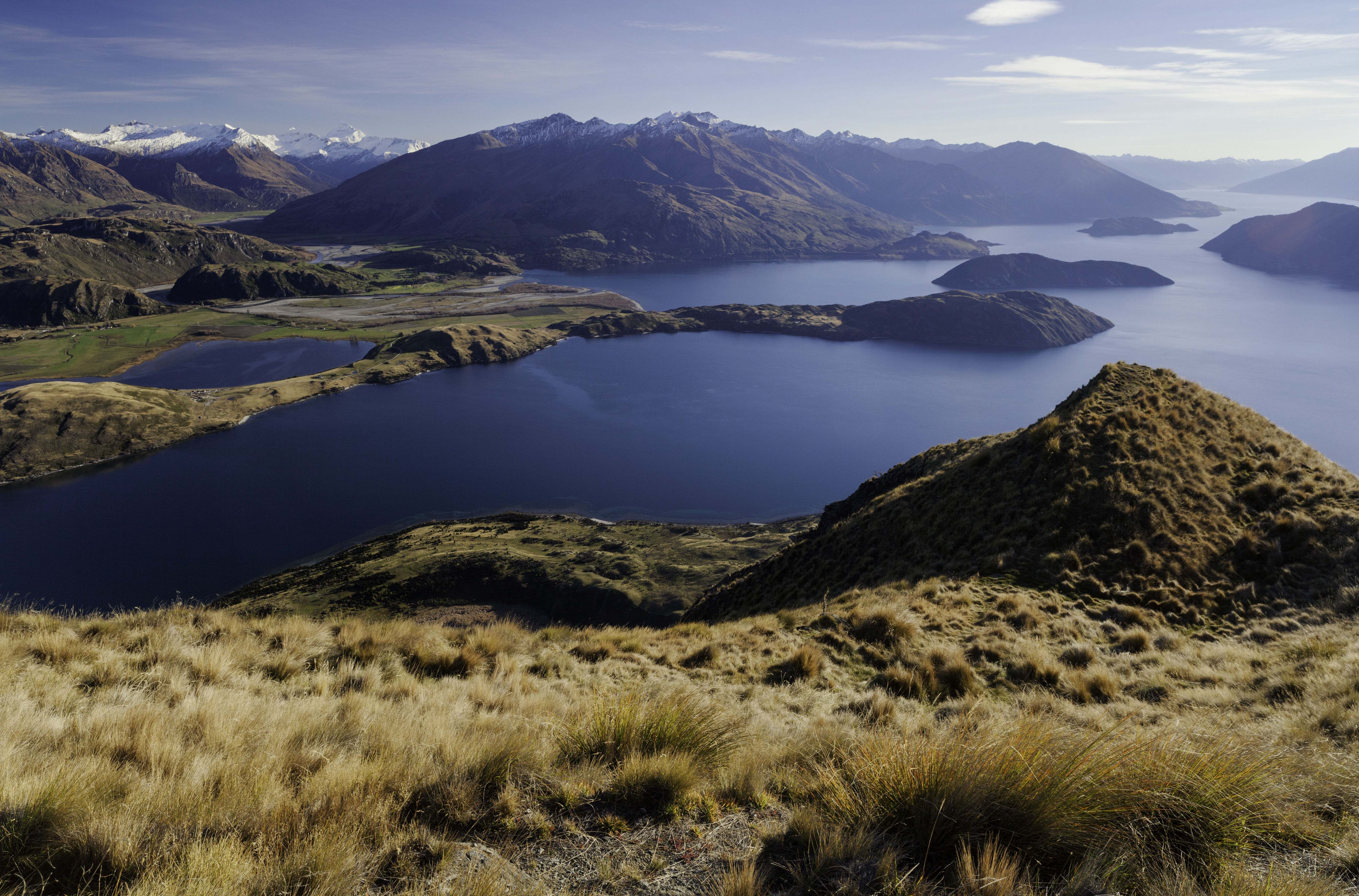 Golden slopes fall toward Lake Wanaka, where blue water and snow-dusted peaks stretch across the distant horizon.
