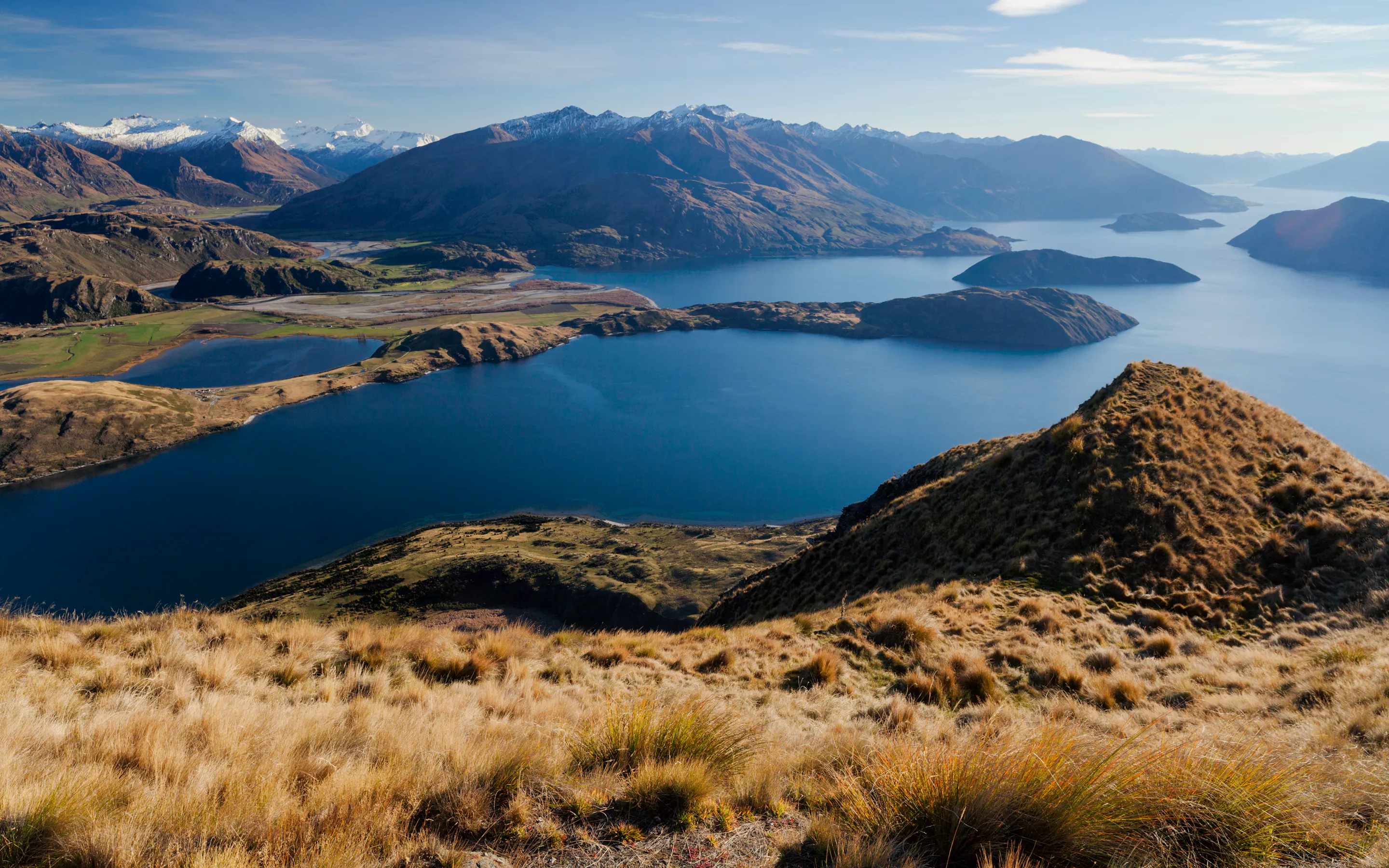 Golden slopes fall toward Lake Wanaka, where blue water and snow-dusted peaks stretch across the distant horizon.