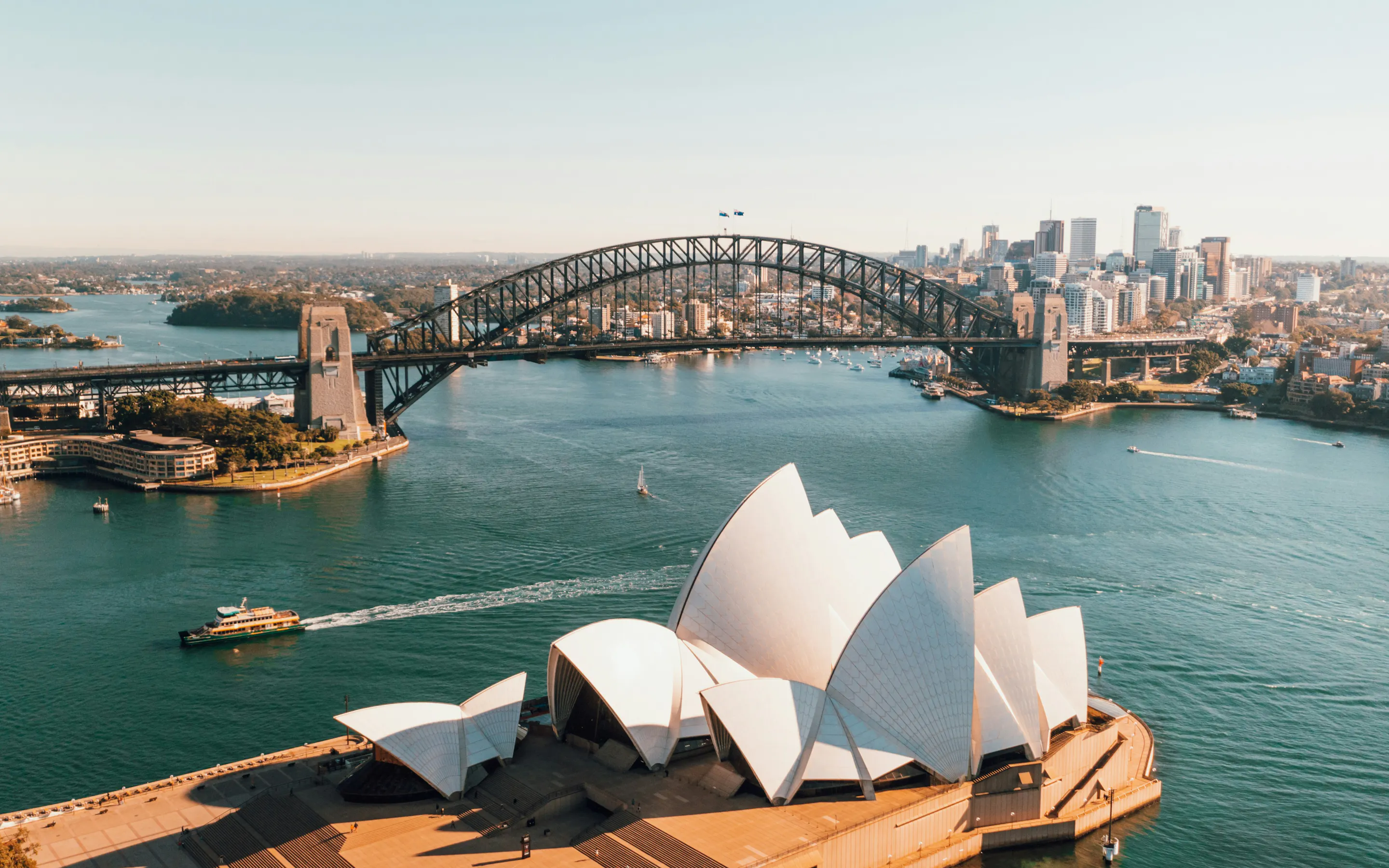 An aerial view frames the Sydney Opera House and Harbour Bridge above blue water, ferries, and the city skyline.