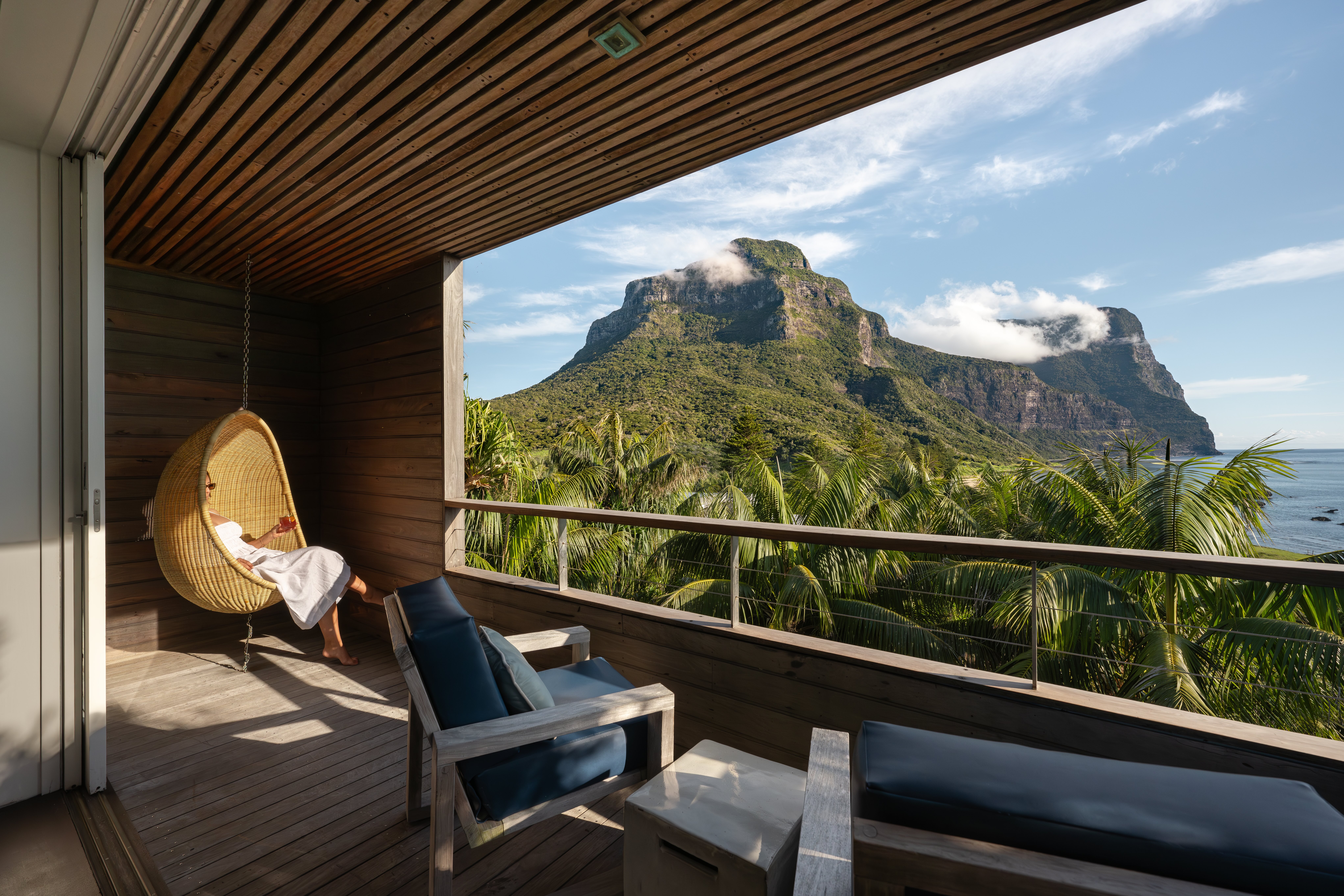 A guest relaxes in a hanging chair on a timber deck, with Capella Lodge palms and Lord Howe peaks beyond.