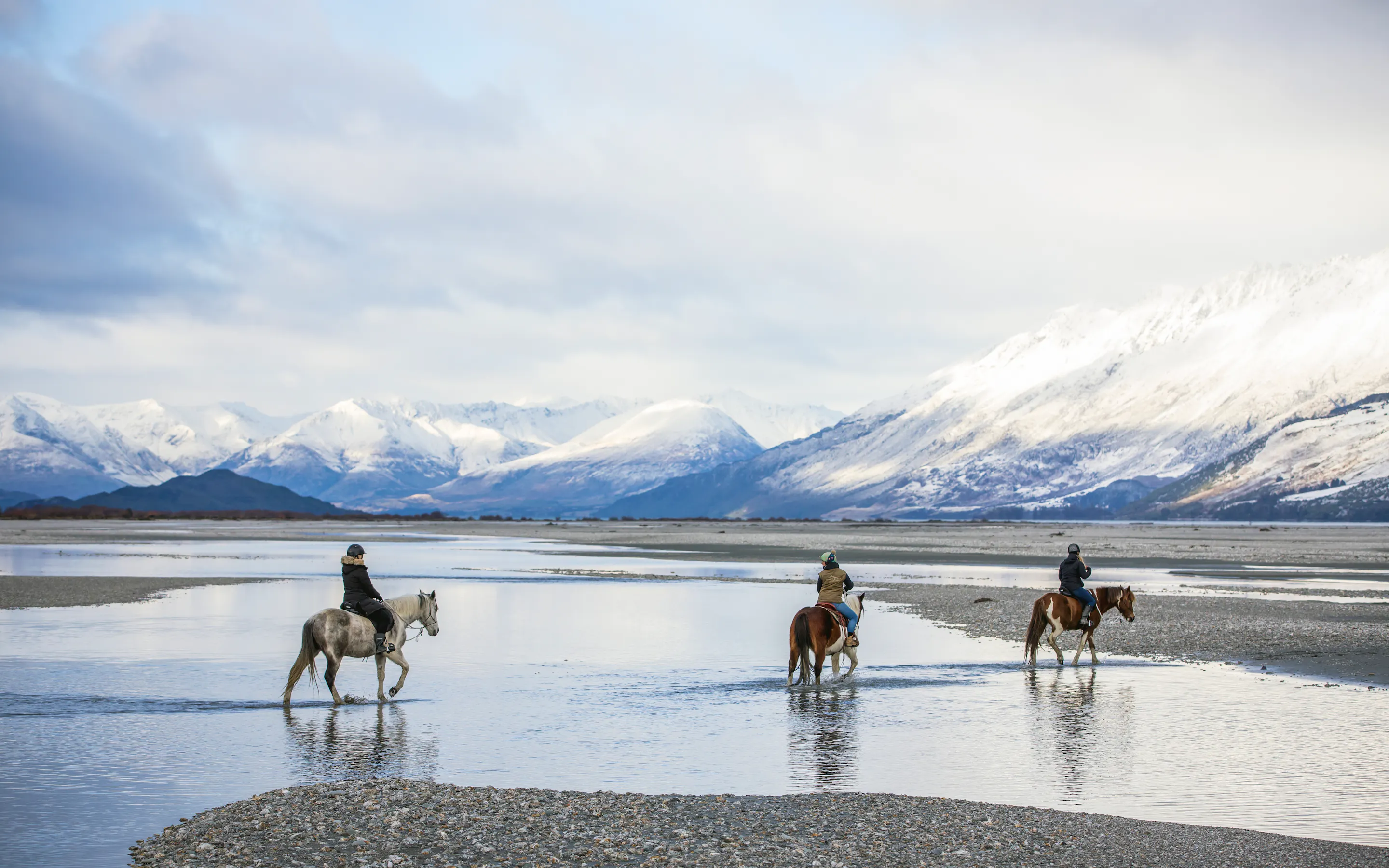 Three riders cross shallow water on horseback with snowy peaks and broad alpine flats stretching behind them.