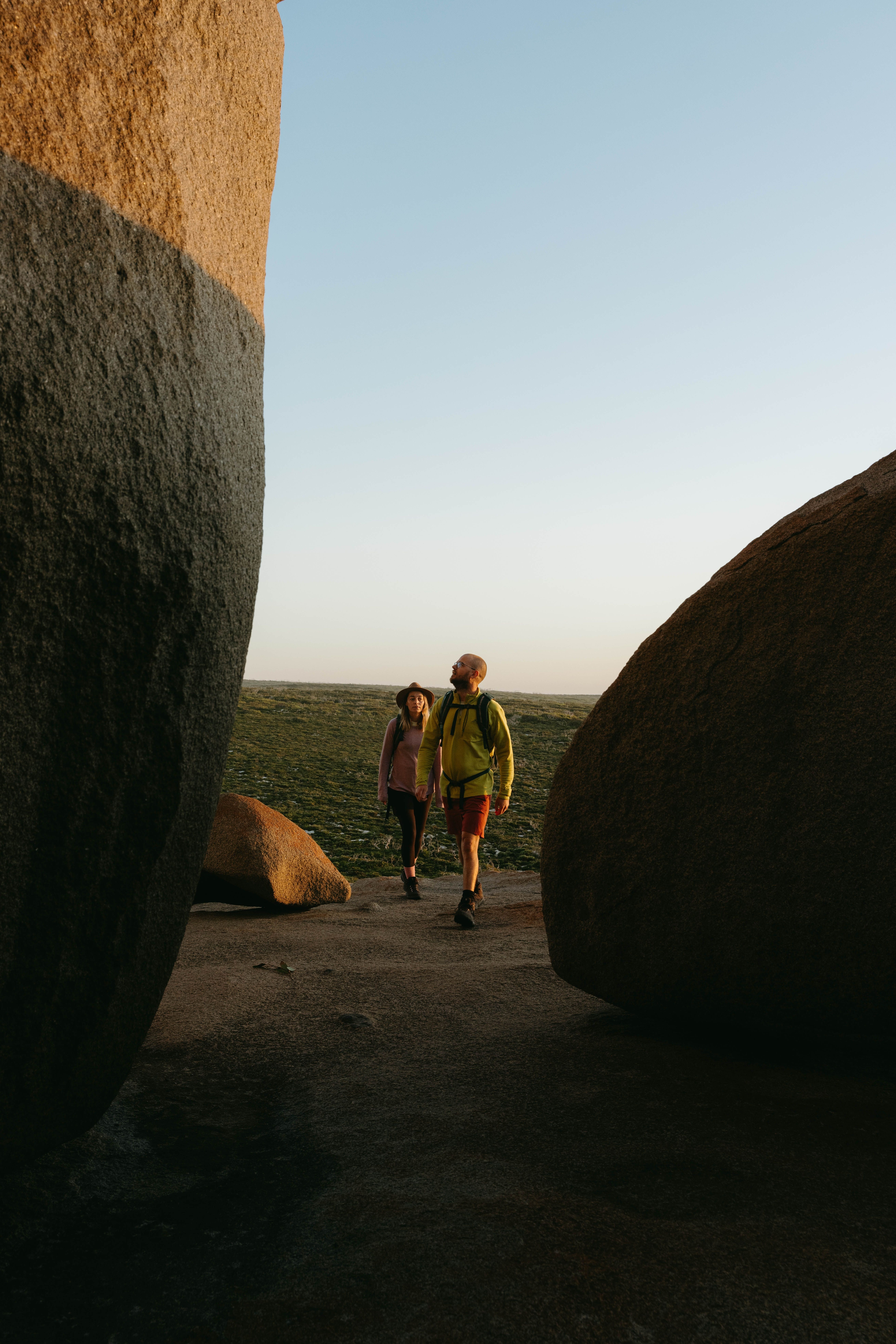 Two hikers walk between rounded granite boulders on a sandy path, with low coastal scrub beyond.