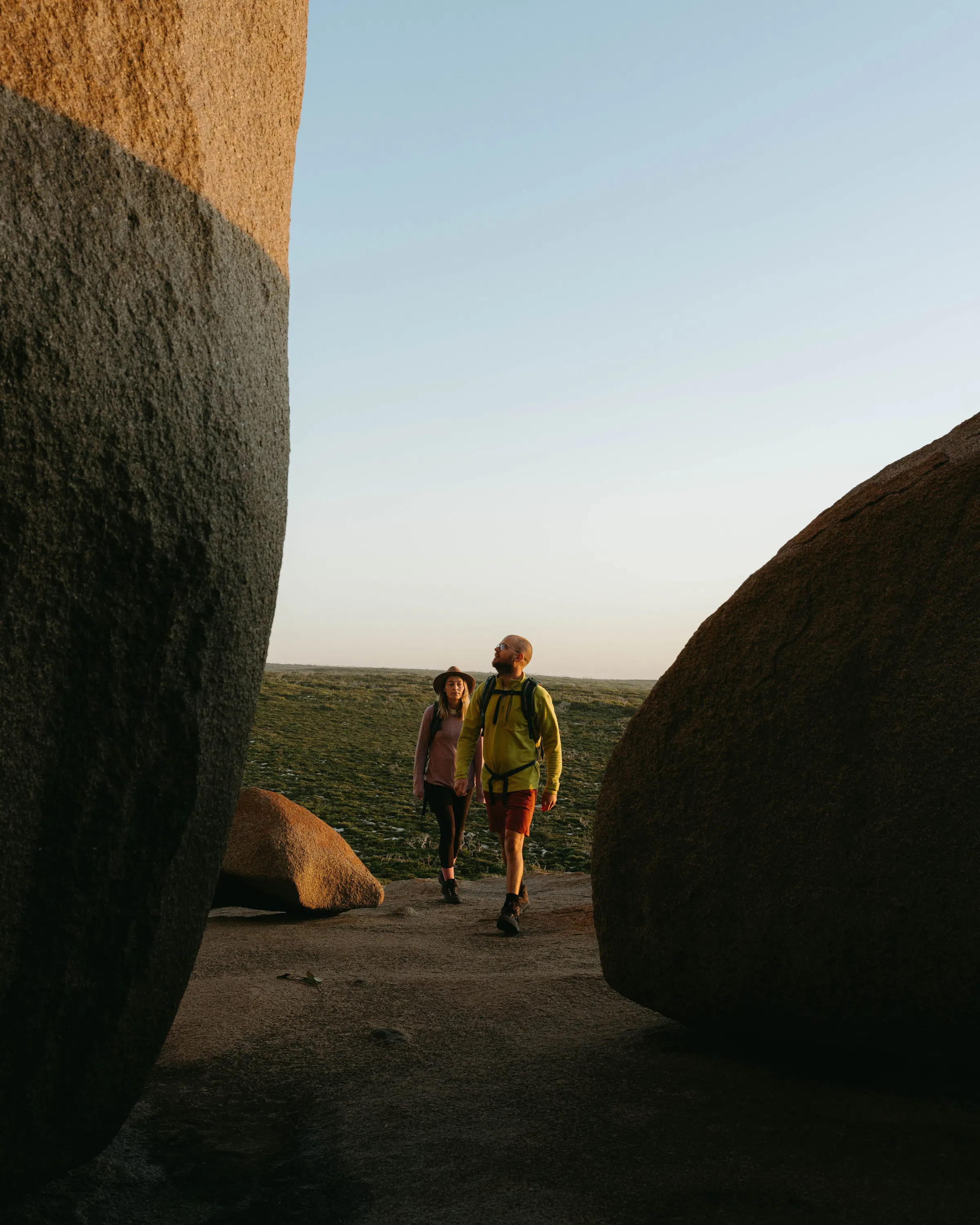 Two hikers walk between rounded granite boulders on a sandy path, with low coastal scrub beyond.