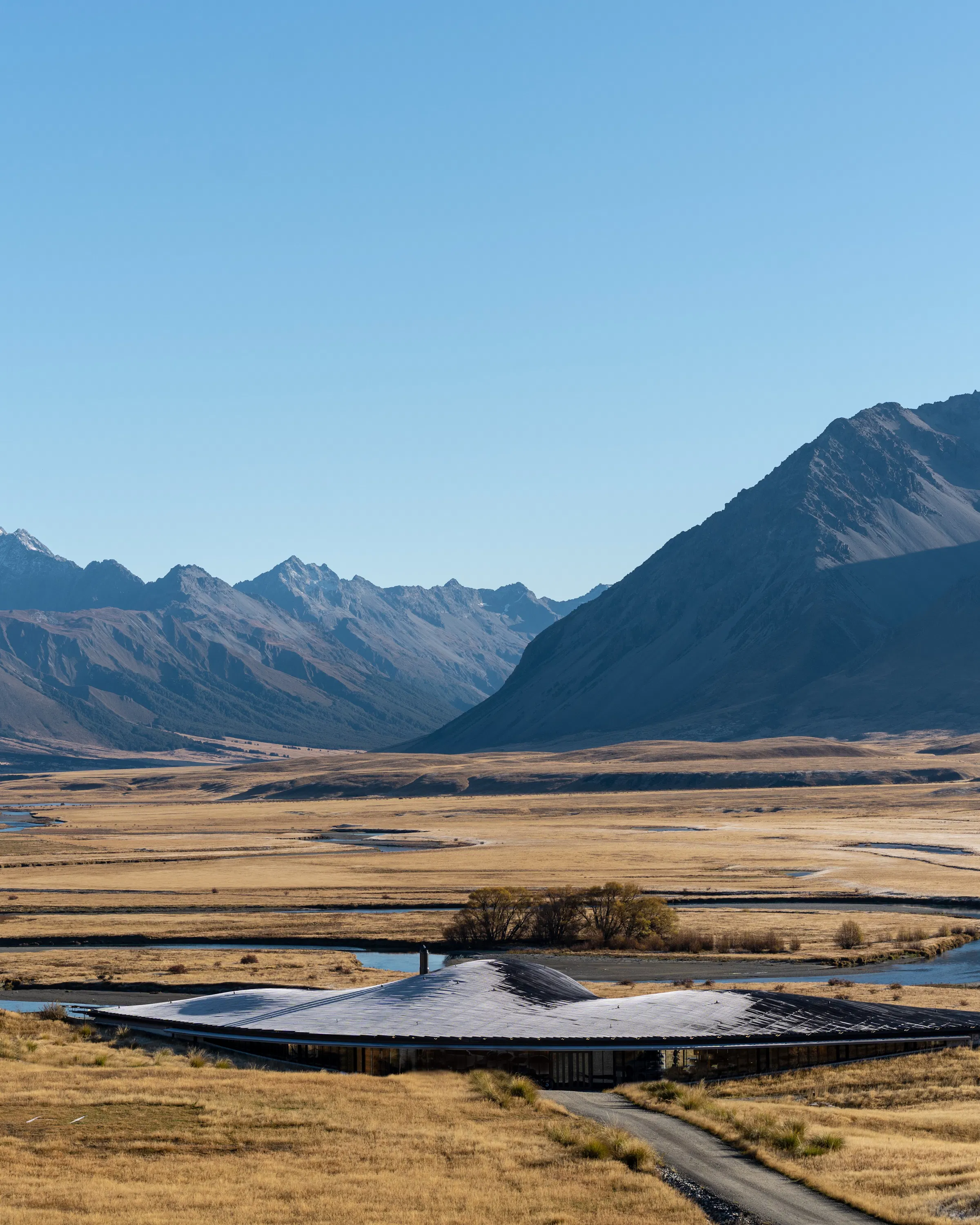 The Lindis Lodge sits low in a broad alpine valley, with braided streams and rugged mountains stretching behind it.