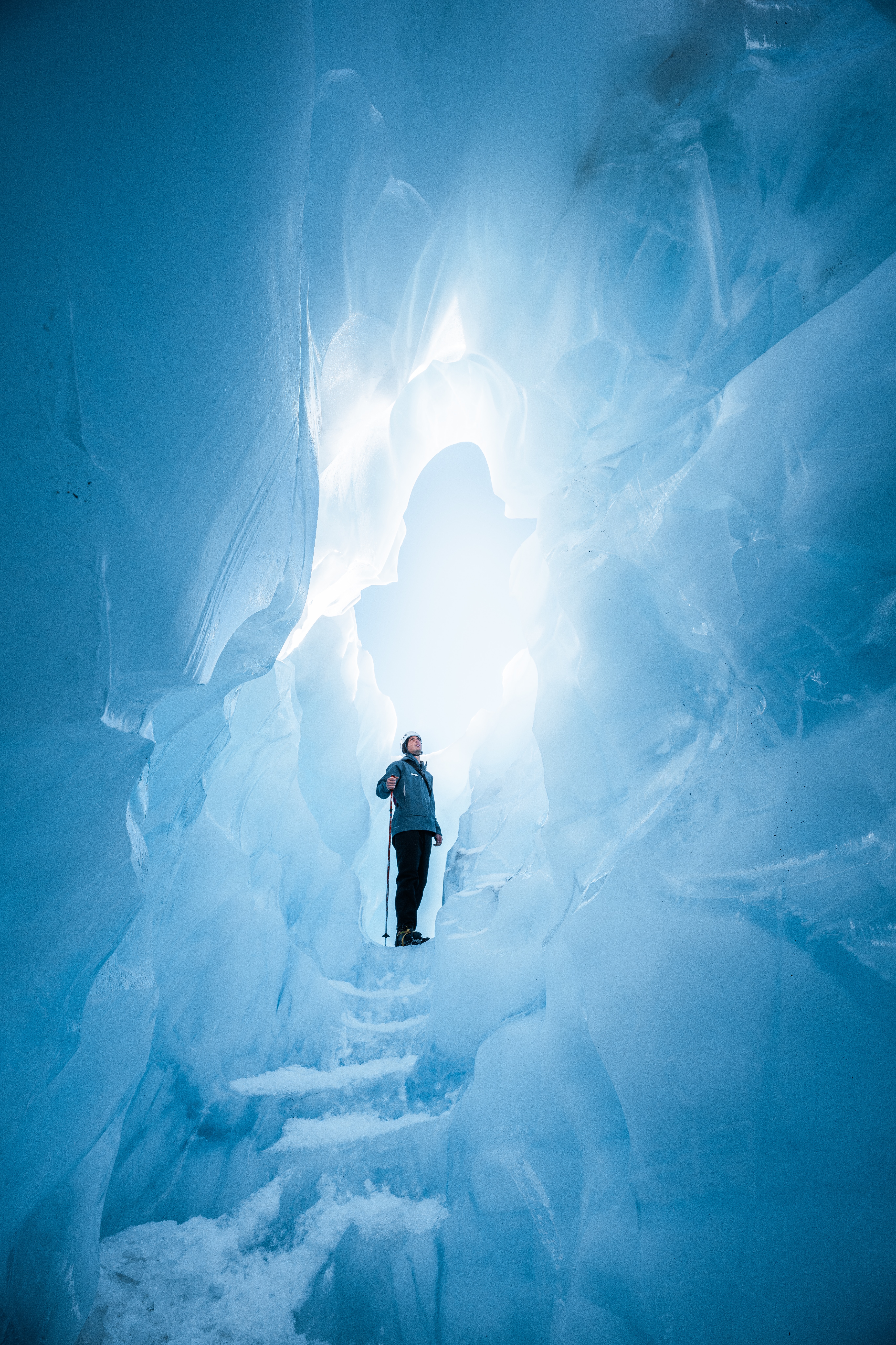 A bundled hiker stands within a blue glacier cave, surrounded by sculpted ice walls and a slick frozen floor.