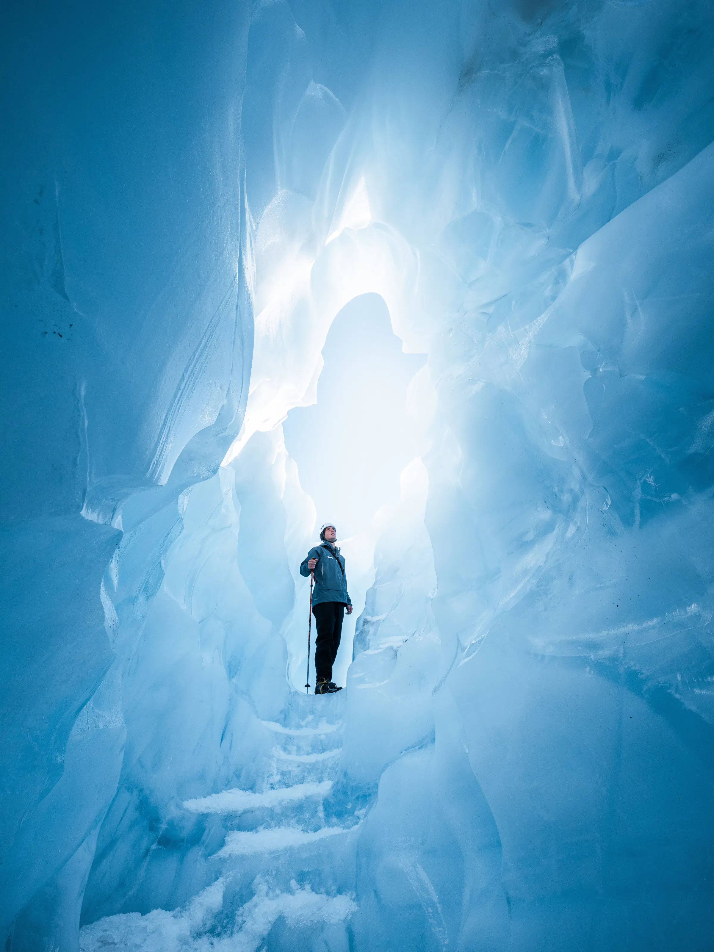 A bundled hiker stands within a blue glacier cave, surrounded by sculpted ice walls and a slick frozen floor.