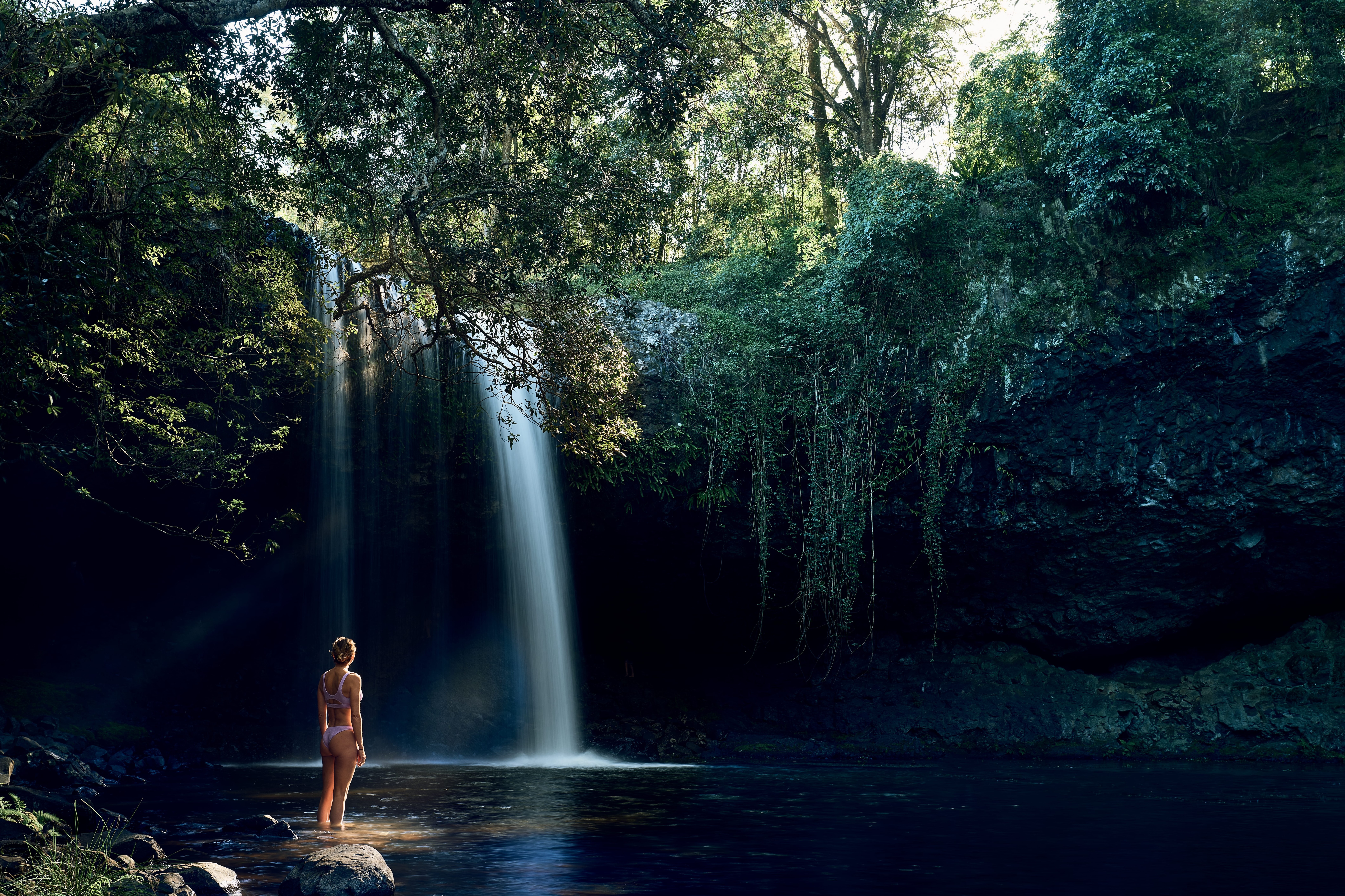 A lone swimmer stands in a dark pool beneath a tall waterfall, framed by overhanging branches and rock walls.