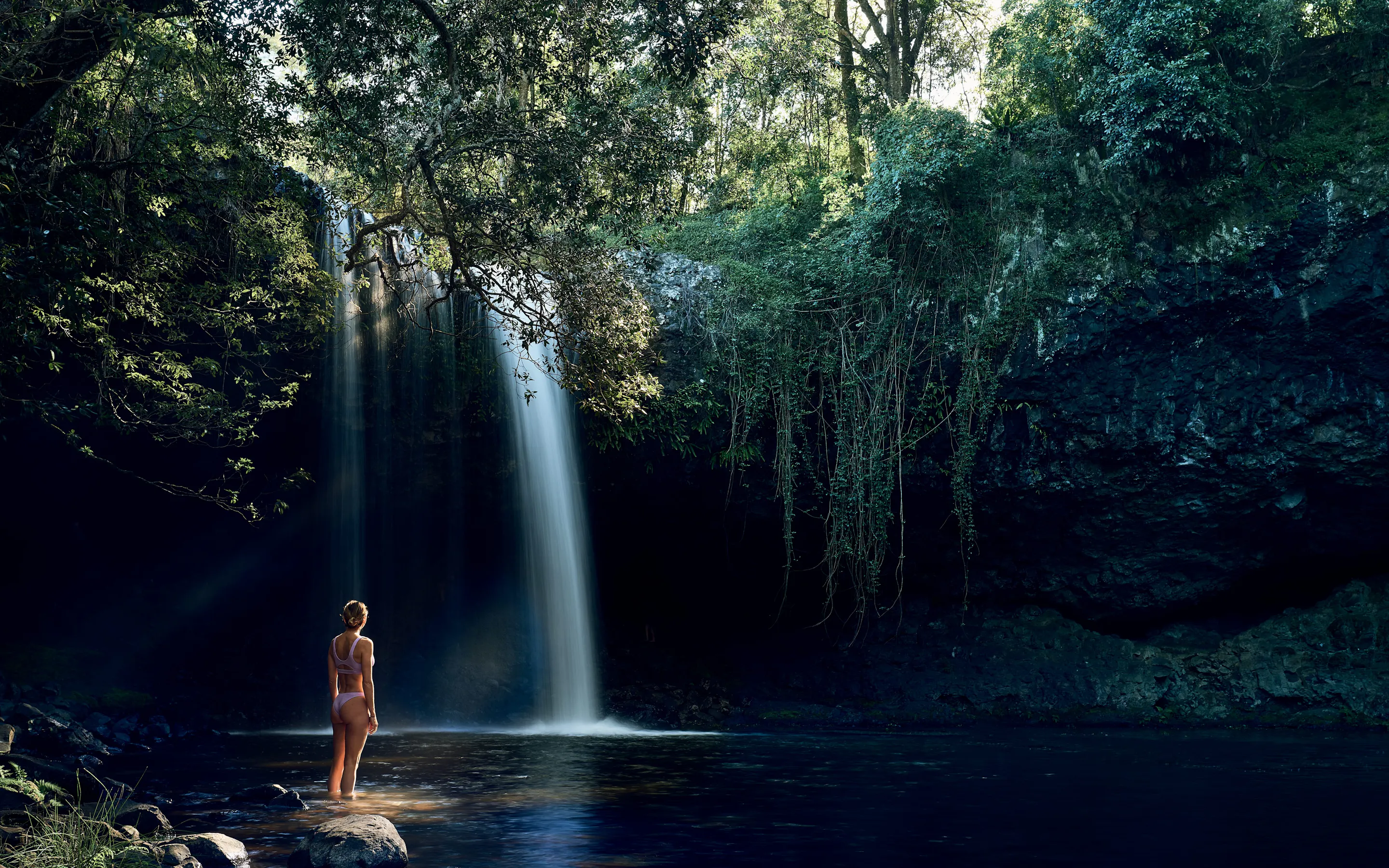 A lone swimmer stands in a dark pool beneath a tall waterfall, framed by overhanging branches and rock walls.