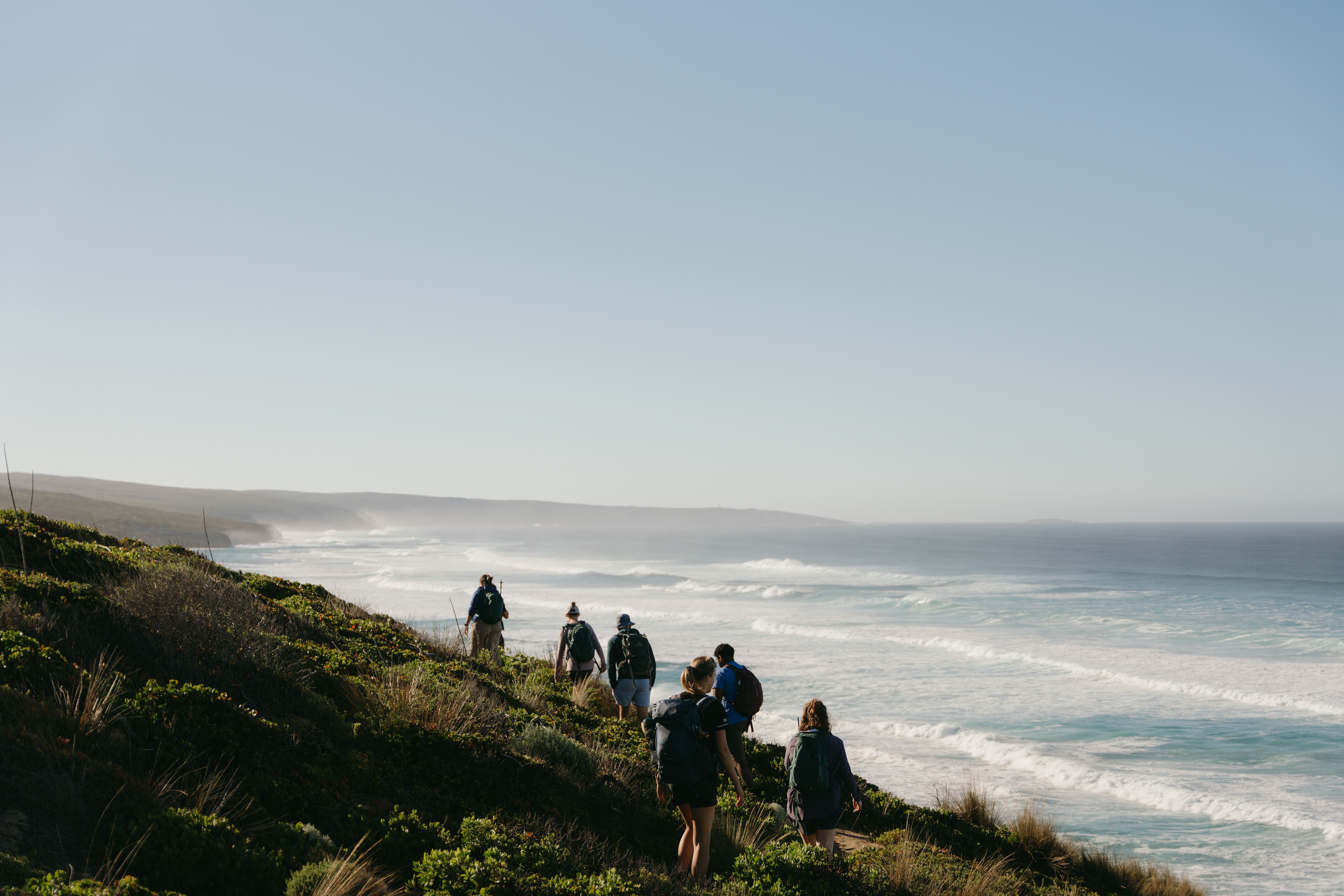 A group of hikers follow a narrow coastal path above rolling surf, with low headlands fading into the hazy distance.