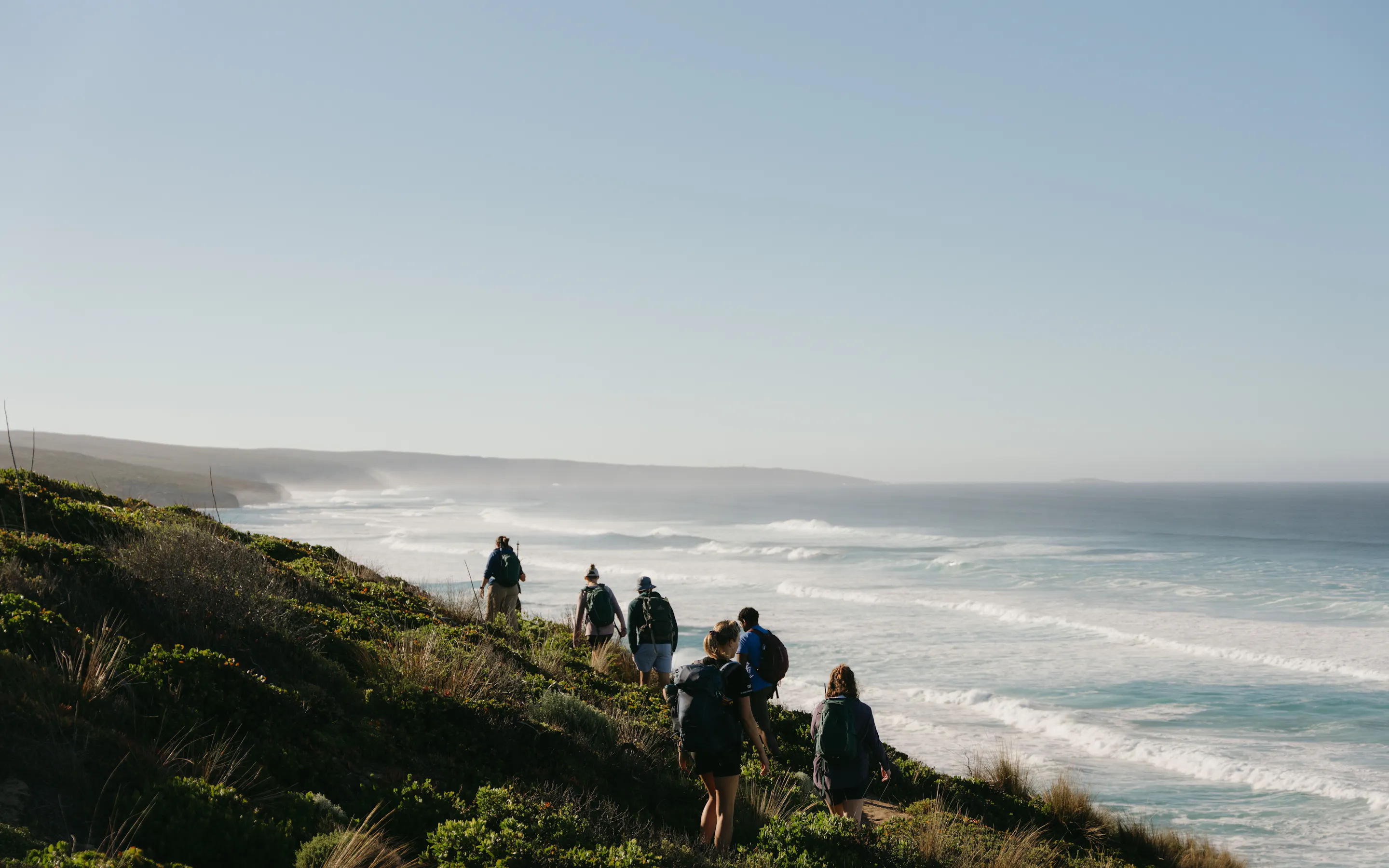 A group of hikers follow a narrow coastal path above rolling surf, with low headlands fading into the hazy distance.
