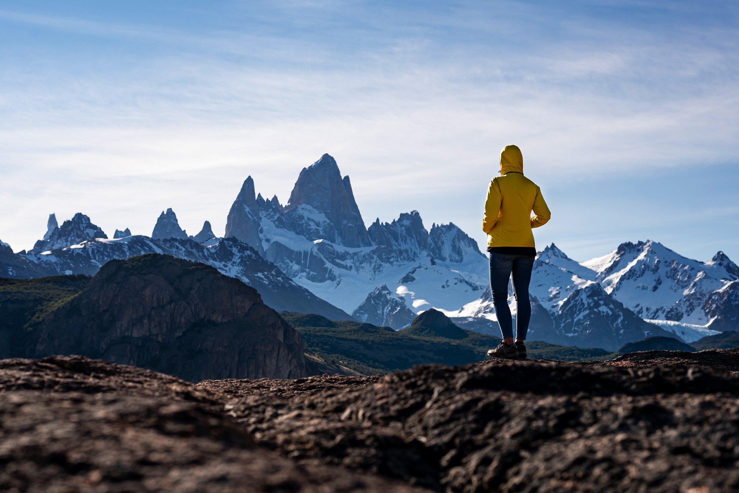 A hiker in a yellow jacket faces the jagged Fitz Roy peaks from a rocky ridge in Argentina's wild Patagonia.