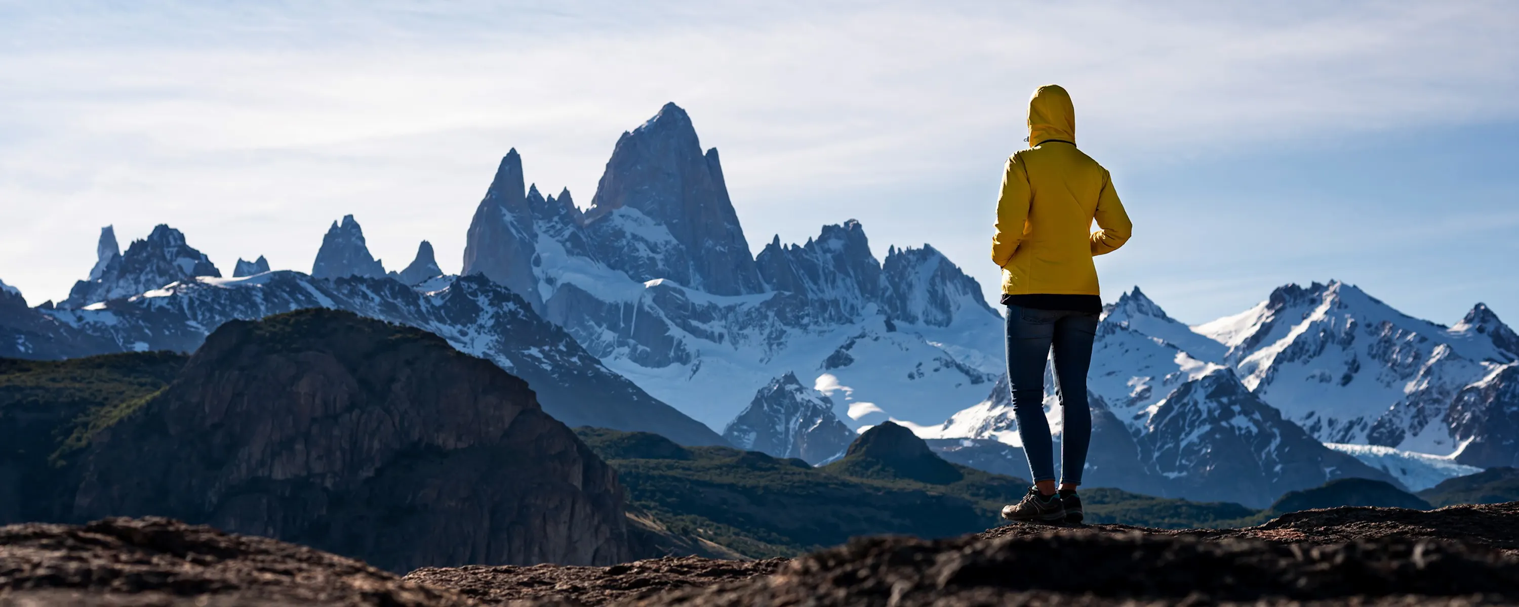 A hiker in a yellow jacket faces the jagged Fitz Roy peaks from a rocky ridge in Argentina's wild Patagonia.
