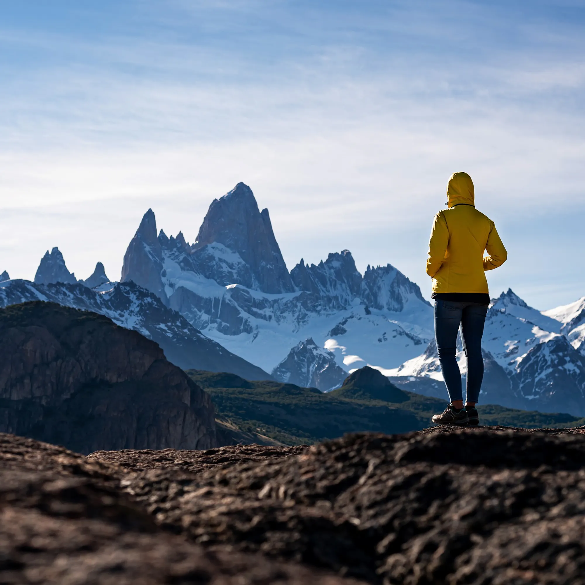 A hiker in a yellow jacket faces the jagged Fitz Roy peaks from a rocky ridge in Argentina's wild Patagonia.