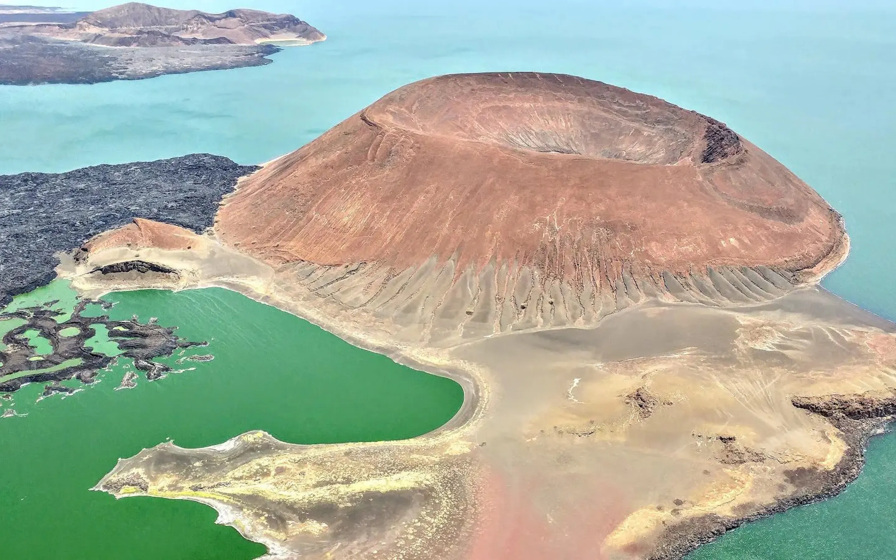 Volcanic Nabuyatom cone rises from Lake Turkana's jade water, with black lava fields and pale shoreline below.