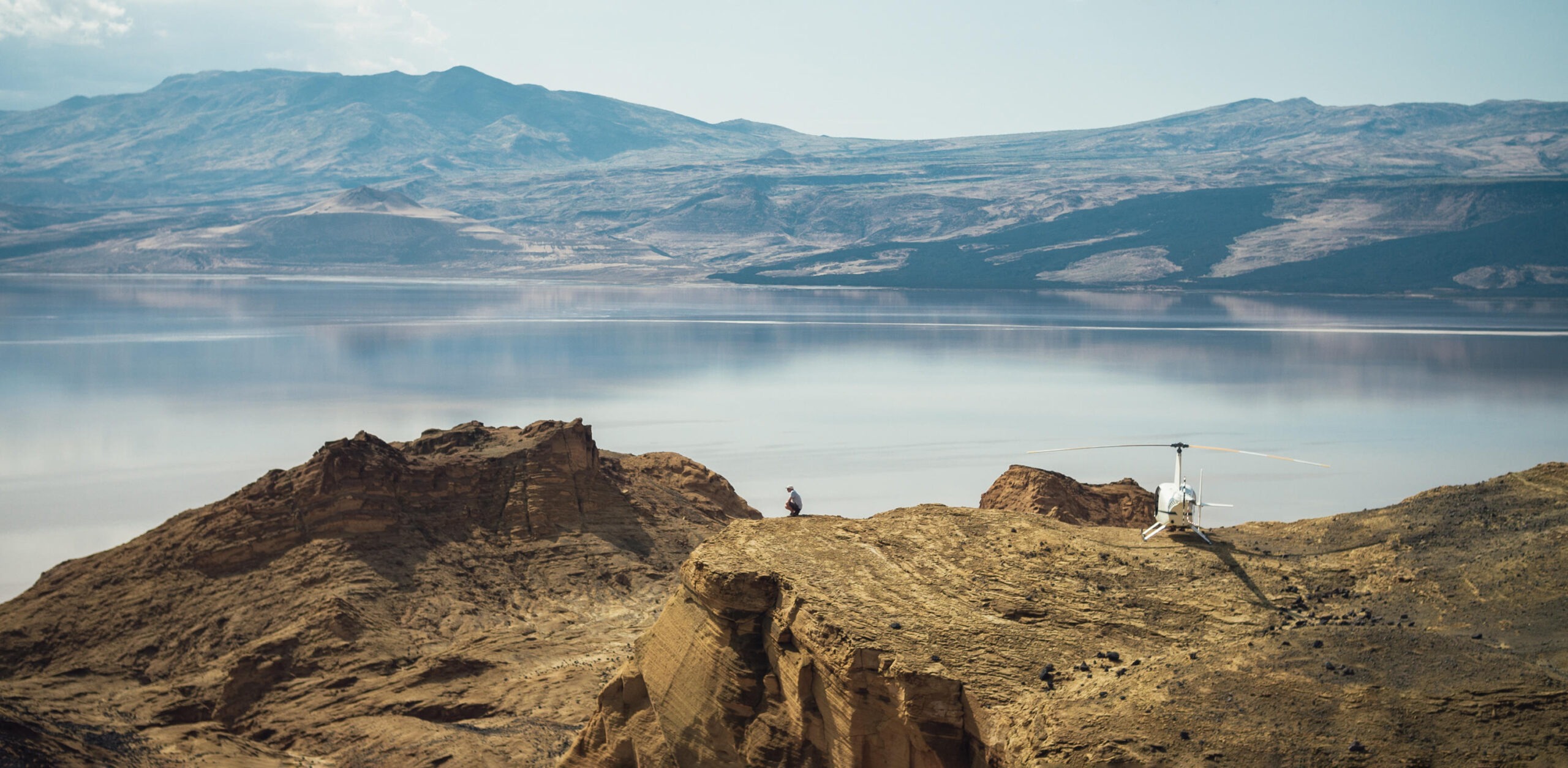 A helicopter rests on a rocky ridge above a calm lake, with hazy mountains reflected across the water in Kenya.
