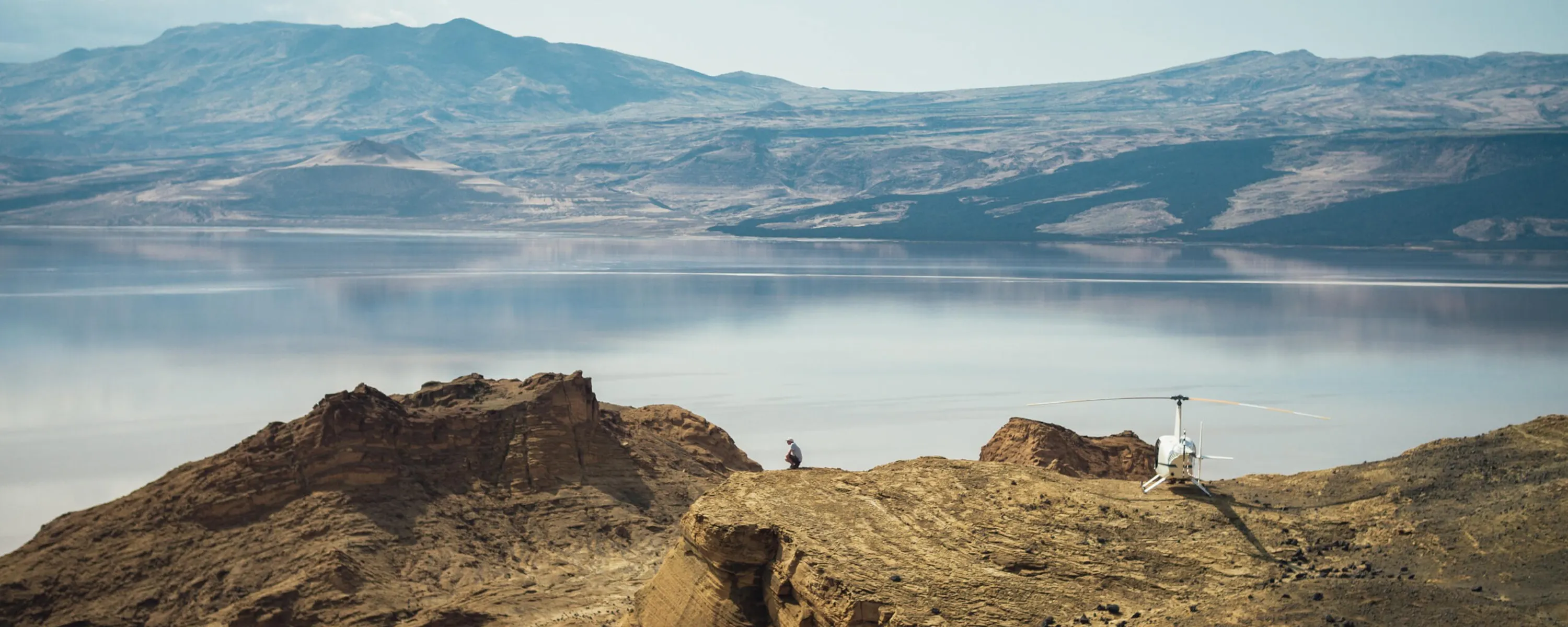 A helicopter rests on a rocky ridge above a calm lake, with hazy mountains reflected across the water in Kenya.