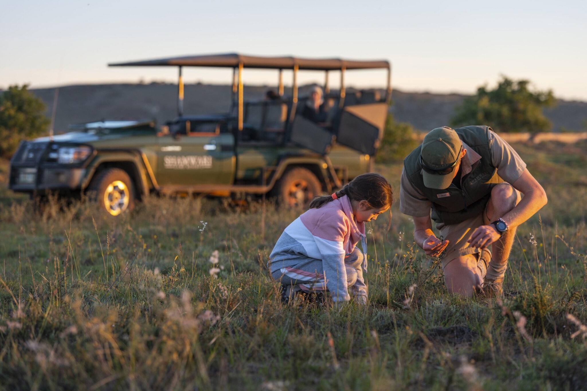 A guide and child kneel in grass near a safari vehicle during a conservation activity at Shamwari in South Africa.