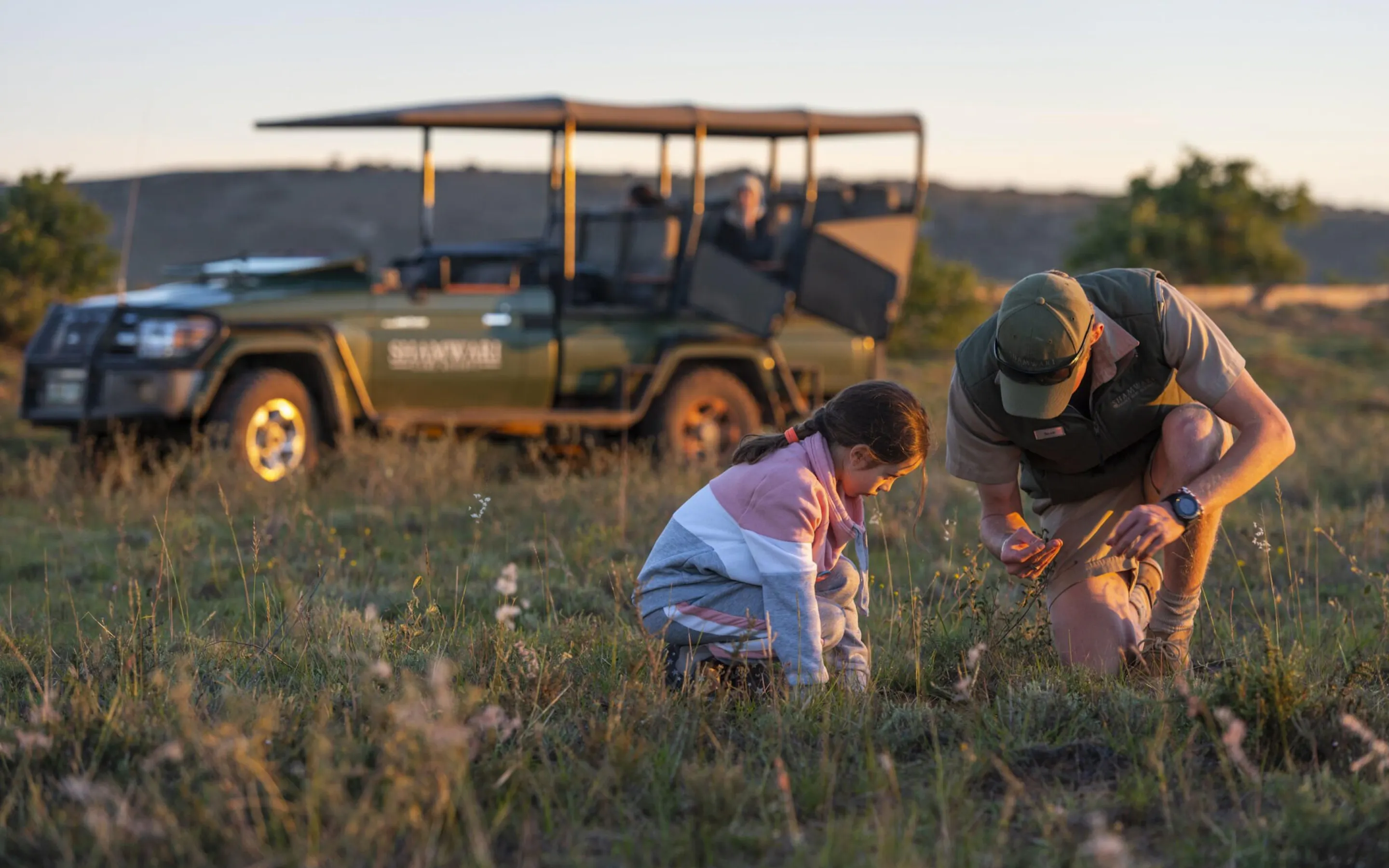 A guide and child kneel in grass near a safari vehicle during a conservation activity at Shamwari in South Africa.