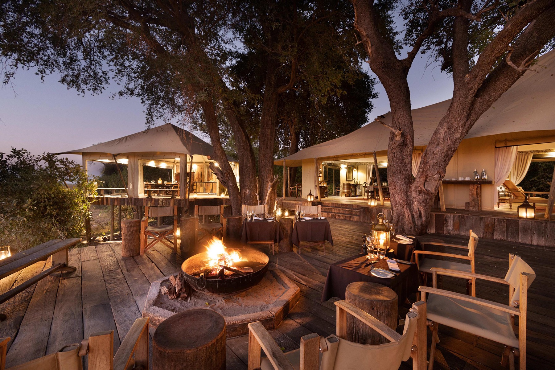 Lanterns, dining tables, and canvas tents surround a glowing fire pit on Zarafa Camp's wooden deck in Botswana.