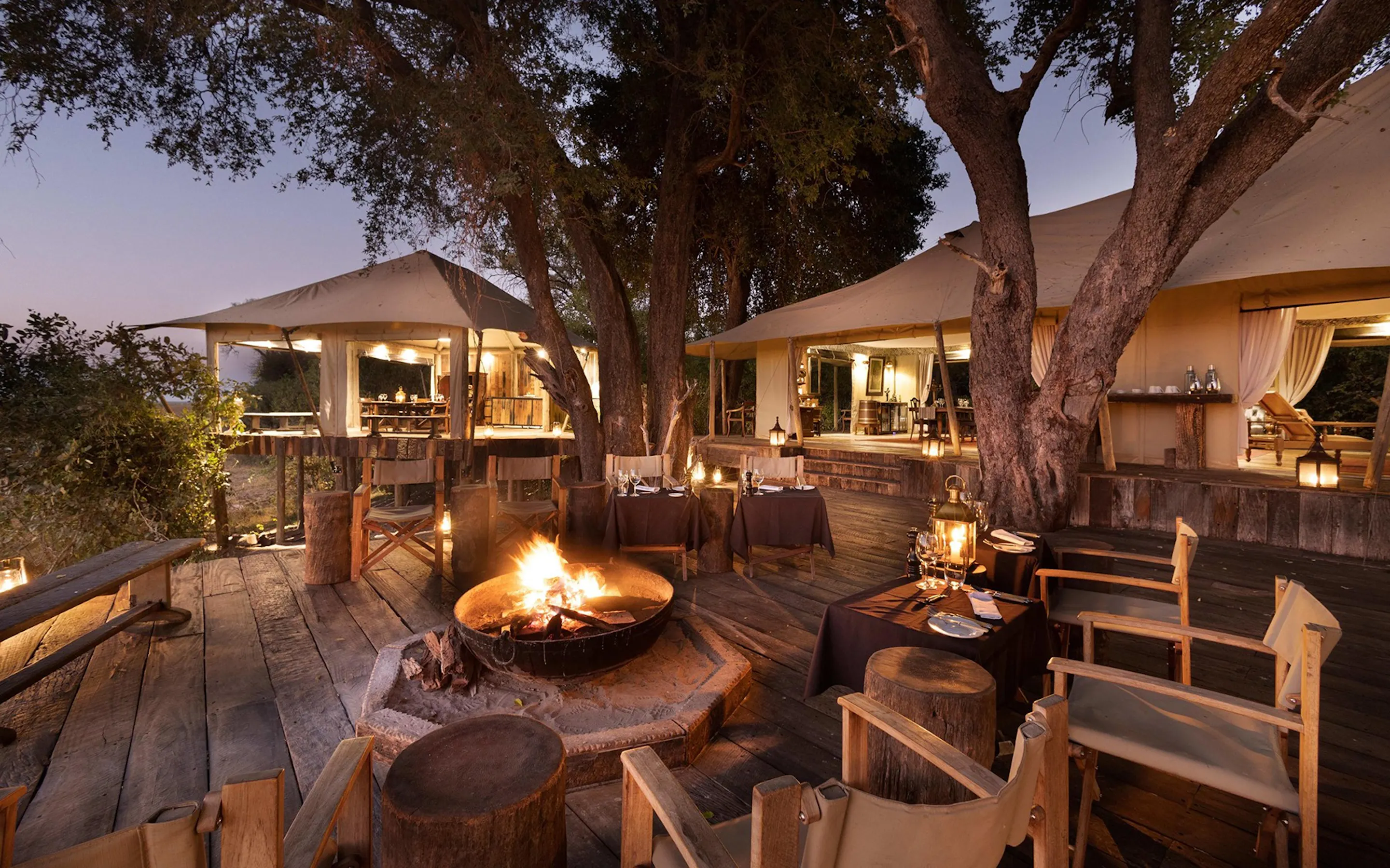Lanterns, dining tables, and canvas tents surround a glowing fire pit on Zarafa Camp's wooden deck in Botswana.
