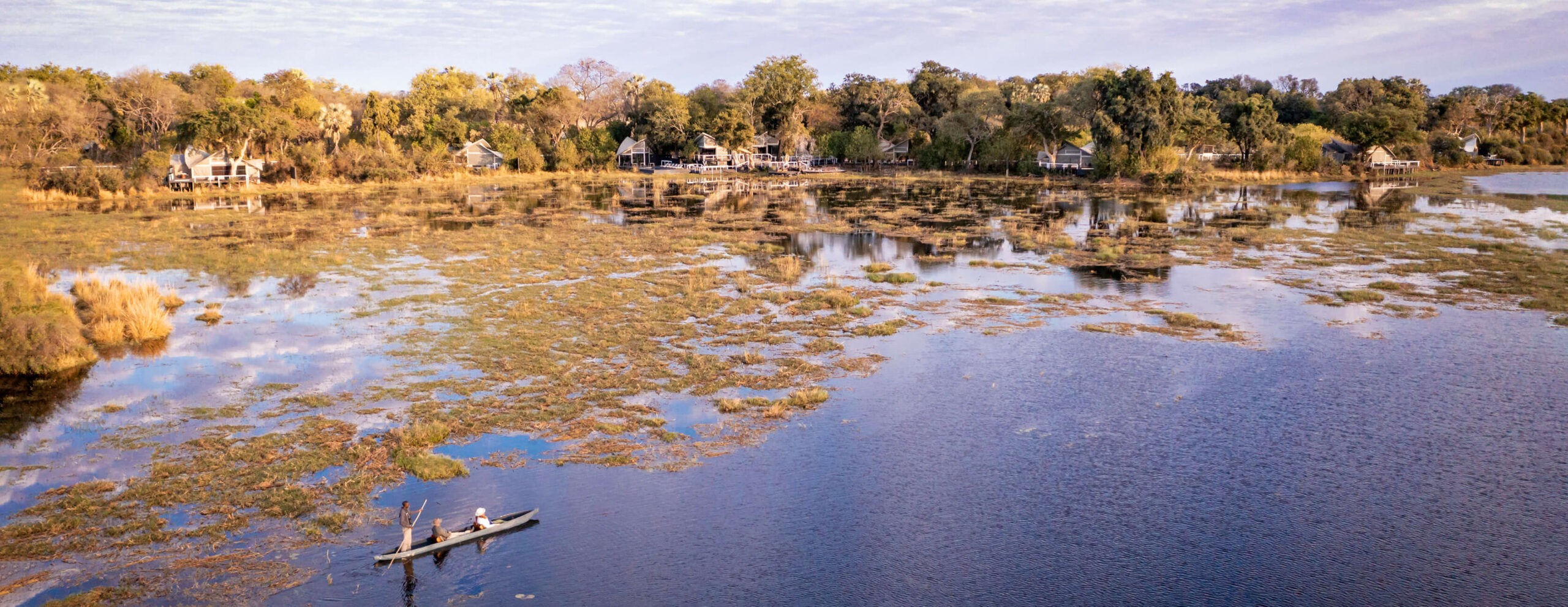 A mokoro glides across shallow water below Abu Camp, where tented suites line the wooded edge of the Okavango.