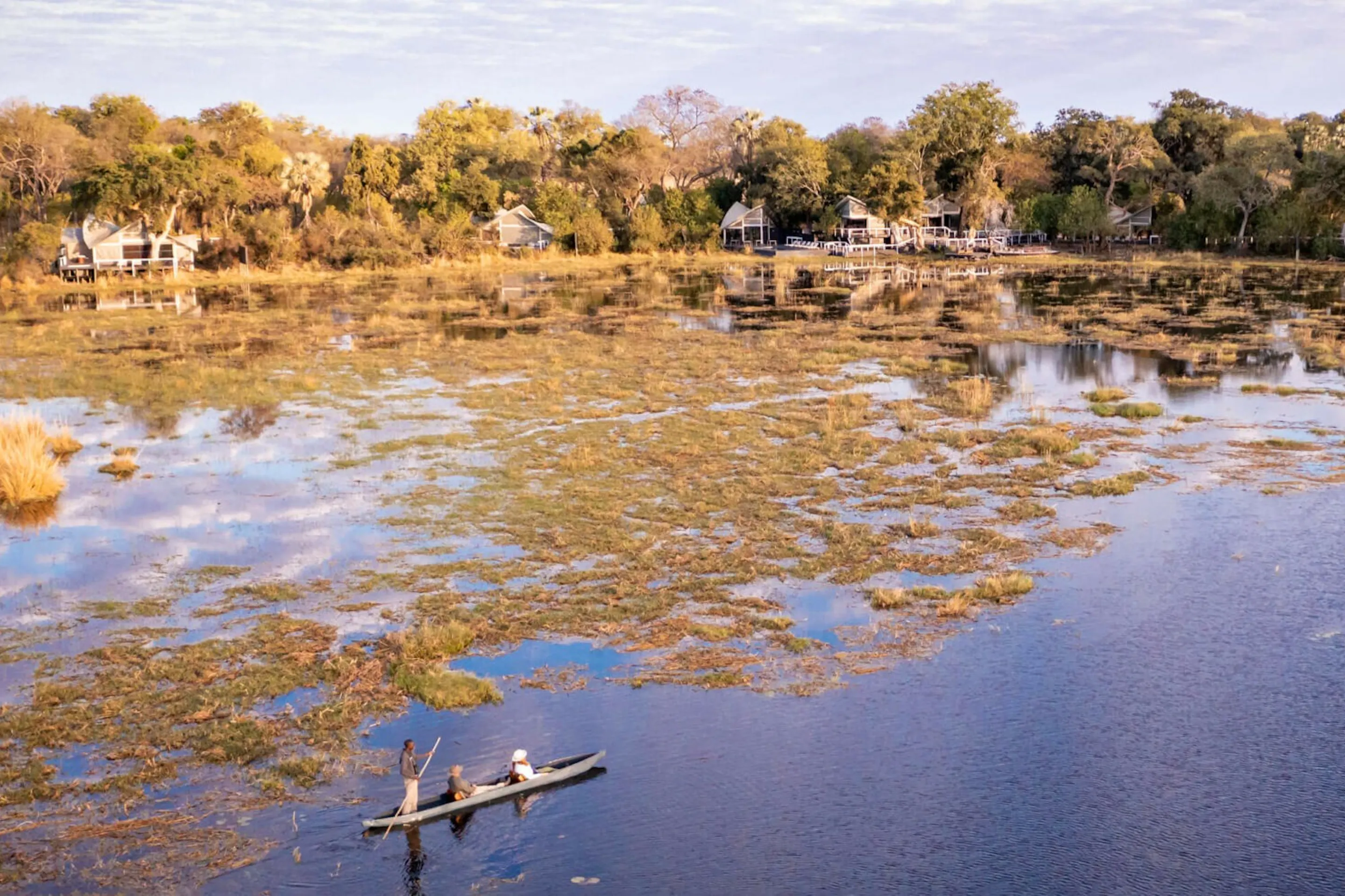 A mokoro glides across shallow water below Abu Camp, where tented suites line the wooded edge of the Okavango.