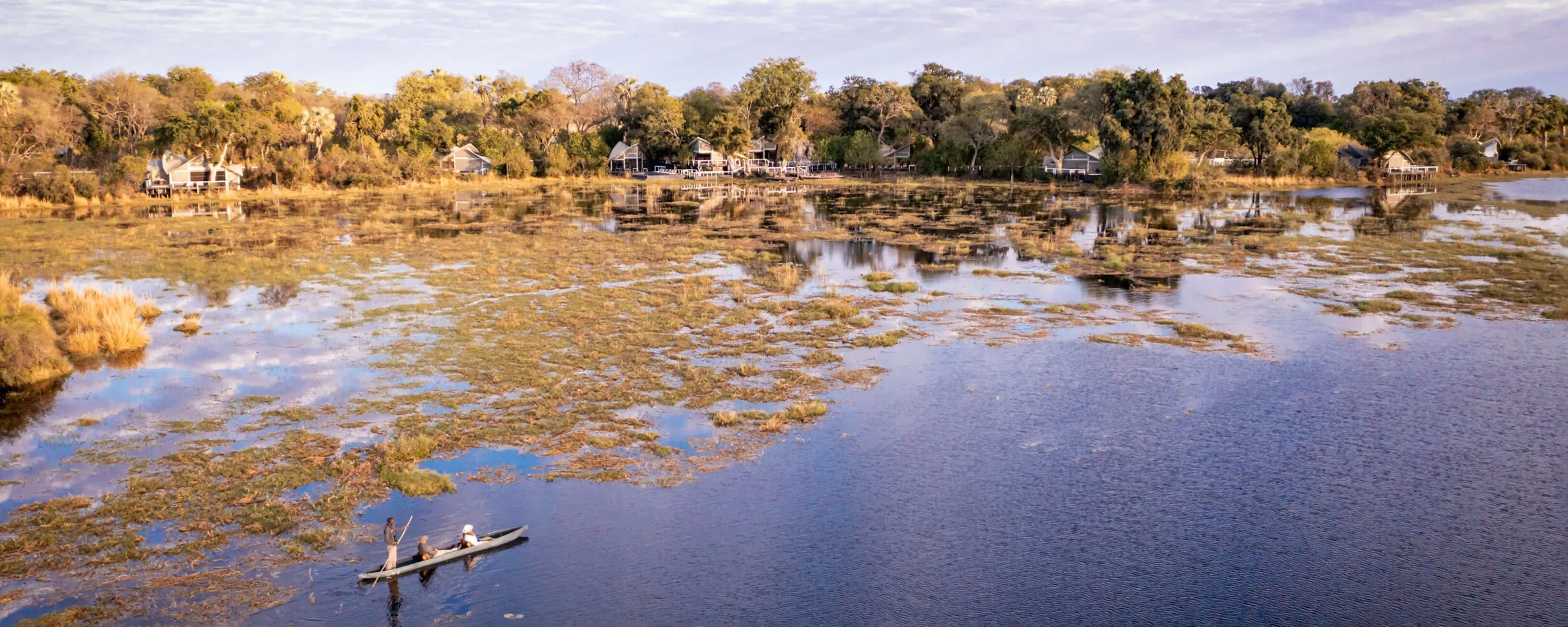 A mokoro glides across shallow water below Abu Camp, where tented suites line the wooded edge of the Okavango.