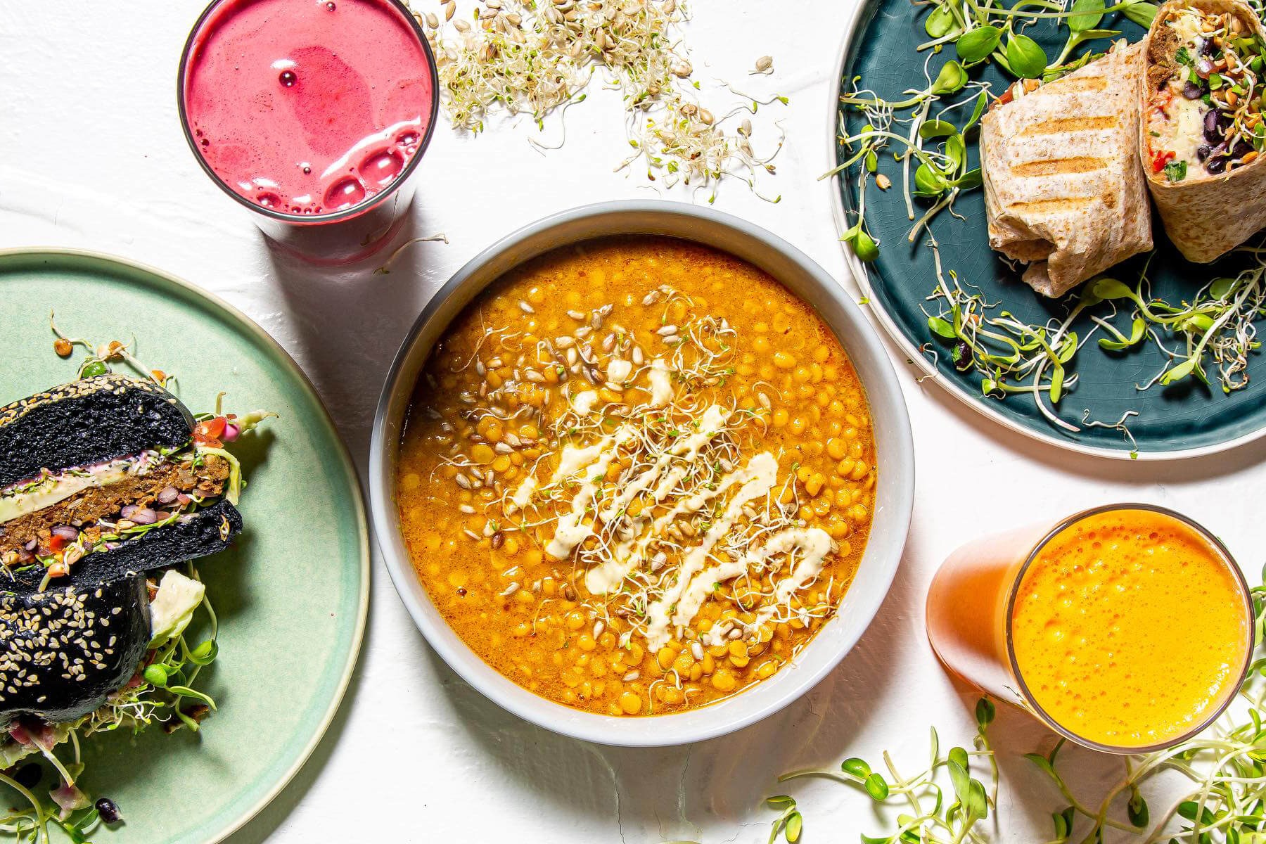 A bright tabletop spread shows black sandwiches, a grilled wrap, orange stew, juices, and sprouts at Makers Landing.