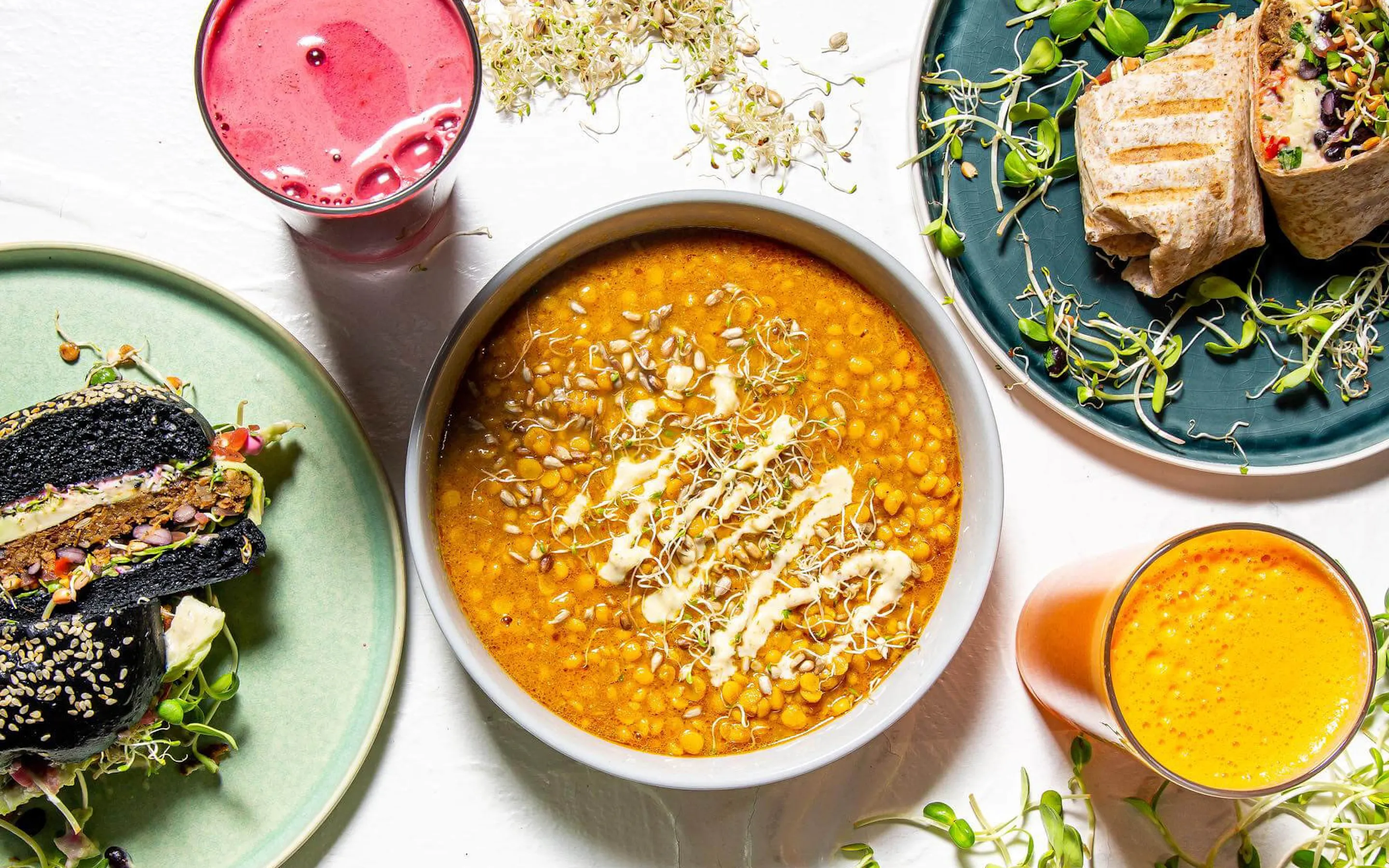 A bright tabletop spread shows black sandwiches, a grilled wrap, orange stew, juices, and sprouts at Makers Landing.