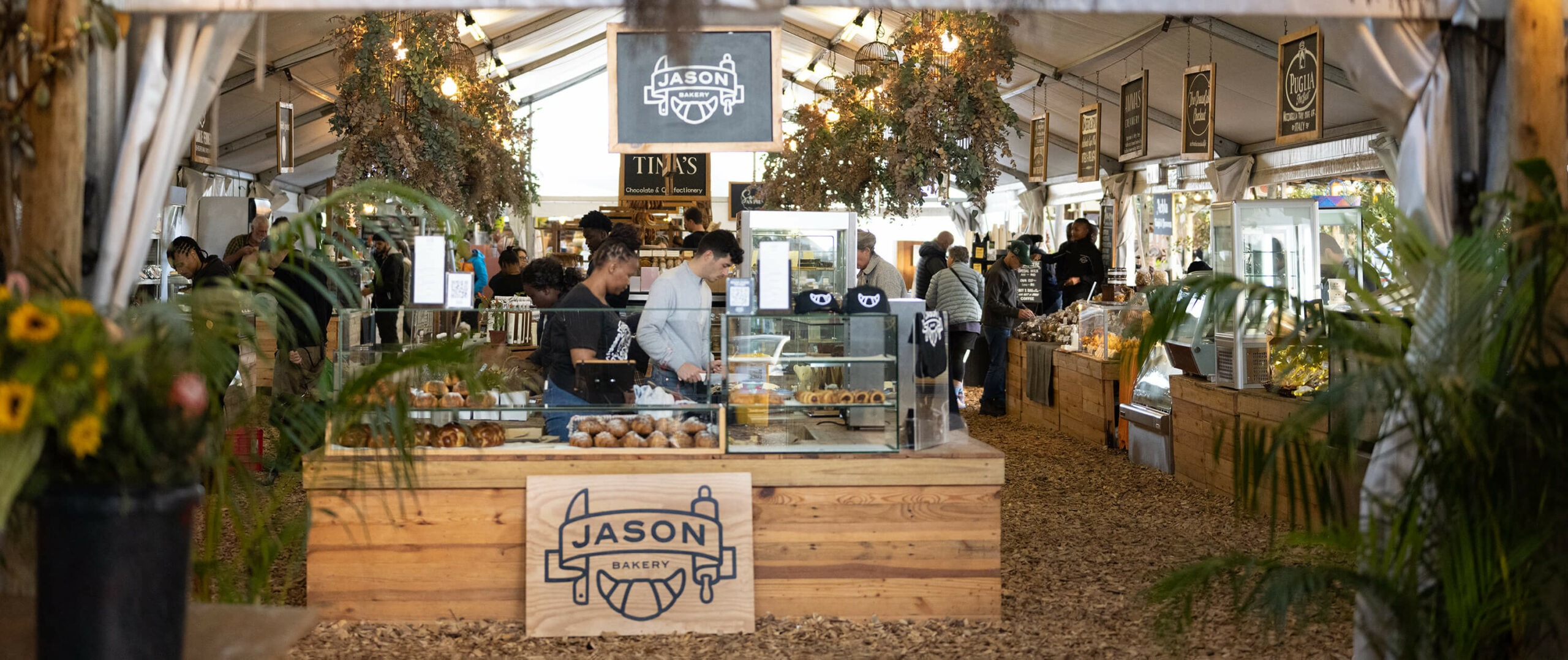A bakery stall with glass pastry cases, wood counters, and hanging signs sits inside a Cape Town market tent.