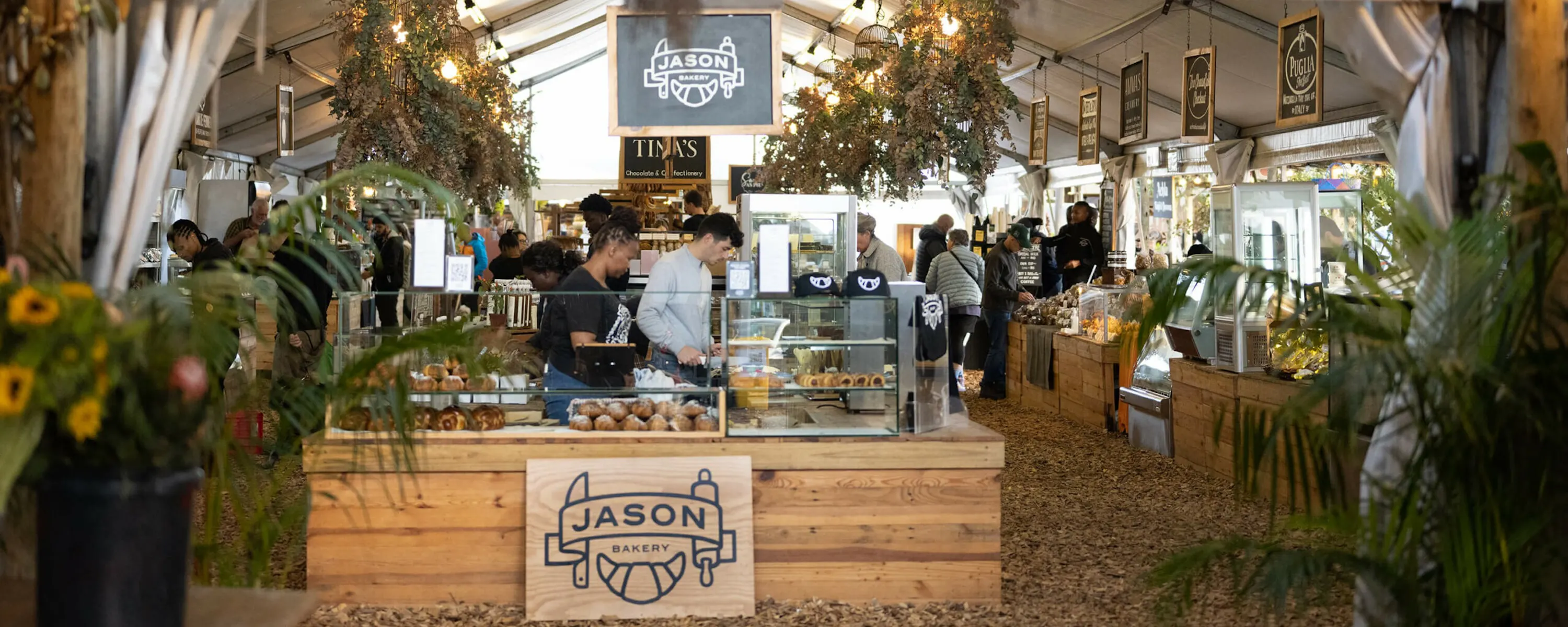 A bakery stall with glass pastry cases, wood counters, and hanging signs sits inside a Cape Town market tent.