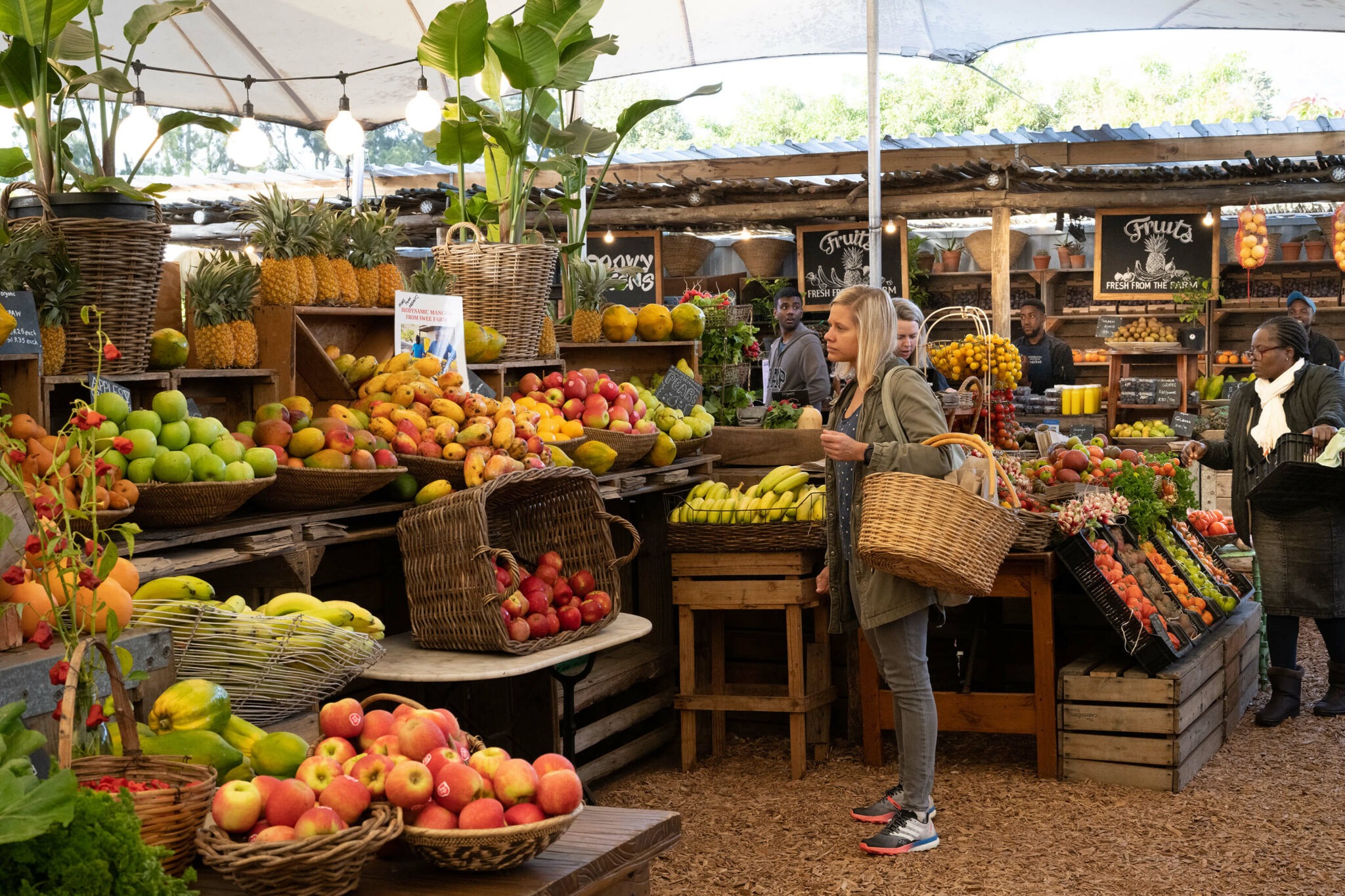 A shopper with a woven basket stands among bright fruit and vegetable displays at Oranjezicht City Farm Market.