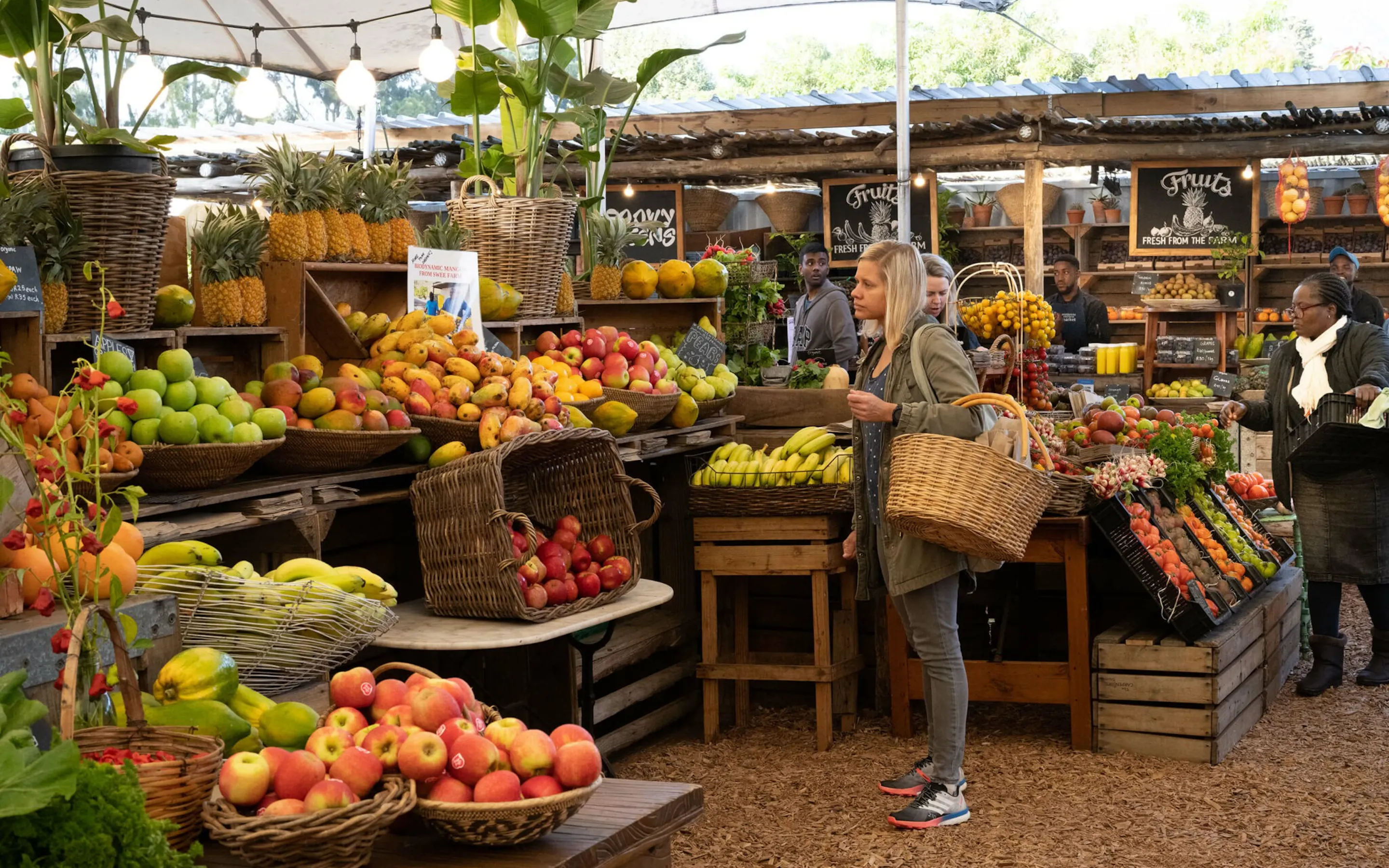 A shopper with a woven basket stands among bright fruit and vegetable displays at Oranjezicht City Farm Market.